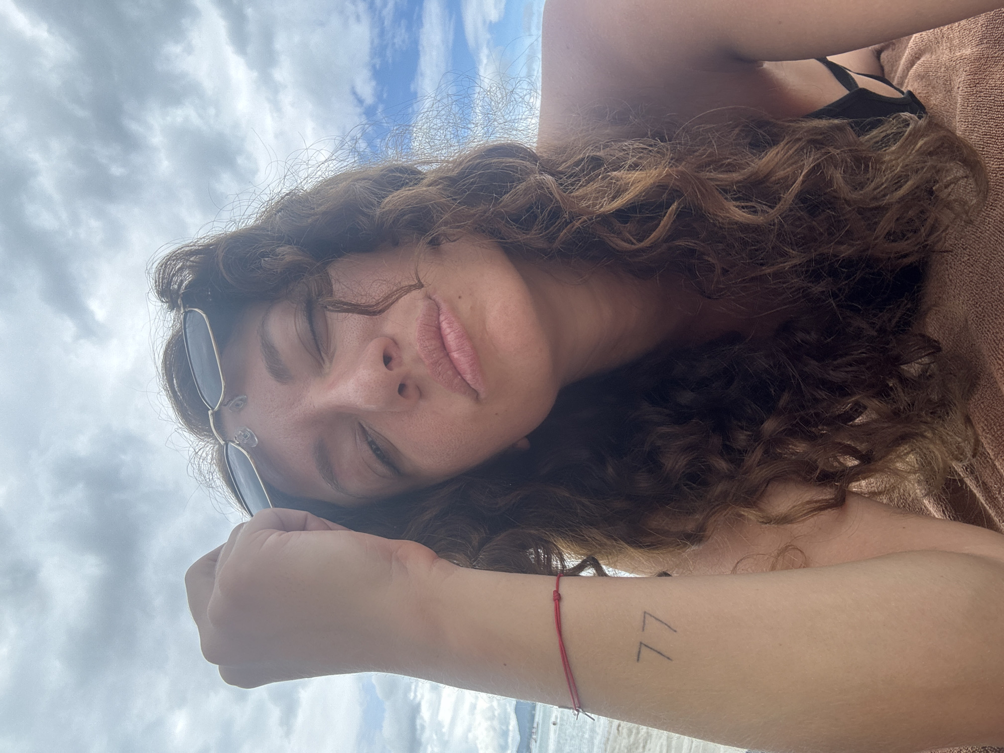 A woman with curly brown hair and sunglasses resting on her head is captured in a close-up selfie on a beach. She is wearing a black bikini top and has a red string bracelet on her left wrist, with the number "77" tattooed on her forearm. Her hand is raised to her forehead, and she is looking at the camera with her eyes partially closed. The background shows a cloudy sky and the ocean with distant islands. The woman is lying on a brown towel. The scene is the beach in the Ko Samui District of Thailand.