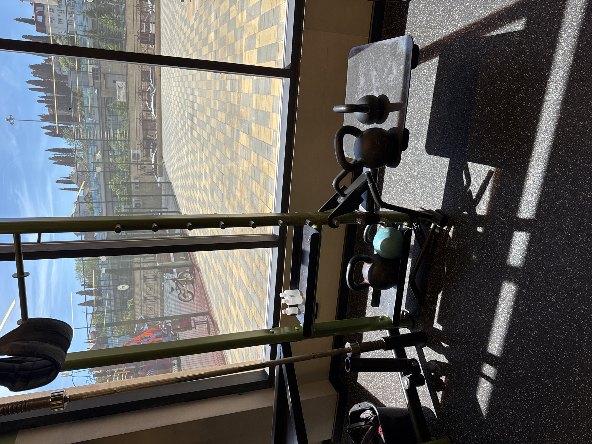 The image captures the interior of a gym looking out through a large window onto an outdoor scene. Inside, various pieces of strength training equipment are visible on a dark, speckled rubberized gym floor. Strong sunlight streams through the window, casting prominent, sharp shadows of the equipment onto the floor, suggesting it is daytime, likely mid-morning or afternoon.
Gym equipment includes several kettlebells, with at least one black and one turquoise visible, alongside a smaller black kettlebell resting on a gray weight bench. A barbell is positioned on a rack that forms part of a robust green metal frame, possibly a squat rack or multi-functional training station. Two small, clear plastic bottles are mounted on the side of this green frame.
The large window offers a clear view of the exterior, which features a distinctive checkered pattern of light and dark yellow or beige paving. Beyond this paved area, there are buildings of varied architecture and numerous tall, slender trees, resembling cypresses. A streetlight and a bicycle are also discernible outside. The sky is bright and blue, indicating clear and sunny weather. No people are visible, and there is no legible text within the frame. The type of vegetation and building styles are consistent with the given location context of Sochi, Russia.