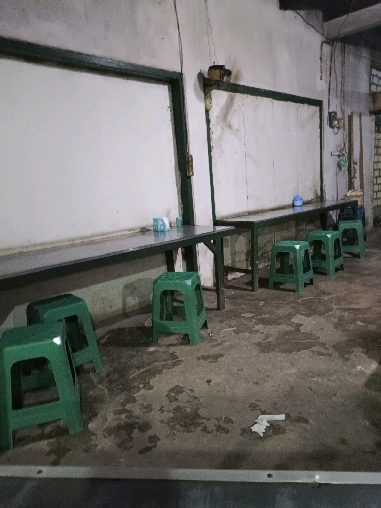 The image depicts an indoor scene, possibly a canteen or a communal eating area, in Sukomulyo, Indonesia. The setting appears utilitarian and somewhat aged, with plain white walls that show signs of wear and staining. A long, dark-colored table runs along the wall, supported by dark green metal legs. Scattered in front of the table are several bright green plastic stools.
Two large, white rectangular boards are mounted on the wall above the table. The board on the left is framed with a dark green border, and the board on the right also has a similar frame. These boards might be used for display, menus, or announcements. On the table in front of the right-hand board, there are a couple of small objects, one appearing to be a light blue container and another a blue and white tissue box. The floor is concrete, with several dark, wet-looking patches, suggesting recent cleaning or spillage.
In the foreground, a white crumpled piece of paper or tissue lies on the floor. Electrical wires are visible along the wall and ceiling, along with some light fixtures. The overall lighting suggests it might be daytime, but the interior is somewhat dimly lit, and there are no windows visible. No people are present in the image, and the scene is still, conveying a sense of quiet or abandonment. The atmosphere is neutral, with no strong emotional cues. There is no visible text in the image.