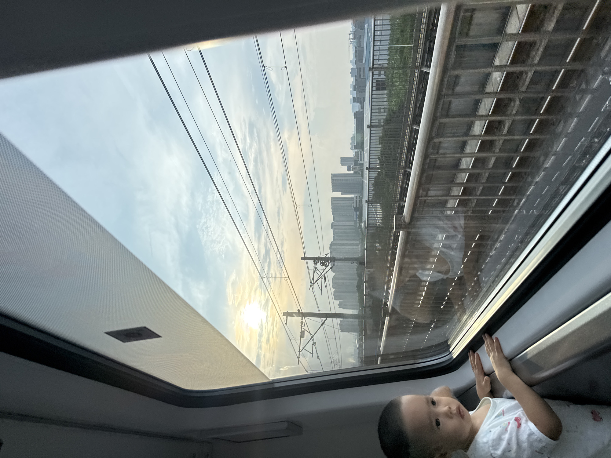 An infant boy with short dark hair, wearing a white t-shirt with a faint pattern, looks out a train window. His hands are resting on the window frame. The train appears to be elevated, with tracks visible below, running parallel to the direction of travel. A metal railing with vertical supports runs alongside the tracks. In the distance, a cityscape with numerous high-rise buildings is visible under a hazy sky with clouds. The sun is low on the horizon, casting a warm glow and creating a lens flare effect. Overhead, power lines and utility poles are strung across the sky. The image seems to be taken from inside the train, looking out. The environment suggests an urban or suburban setting in Nanhai District, China, during either sunrise or sunset.