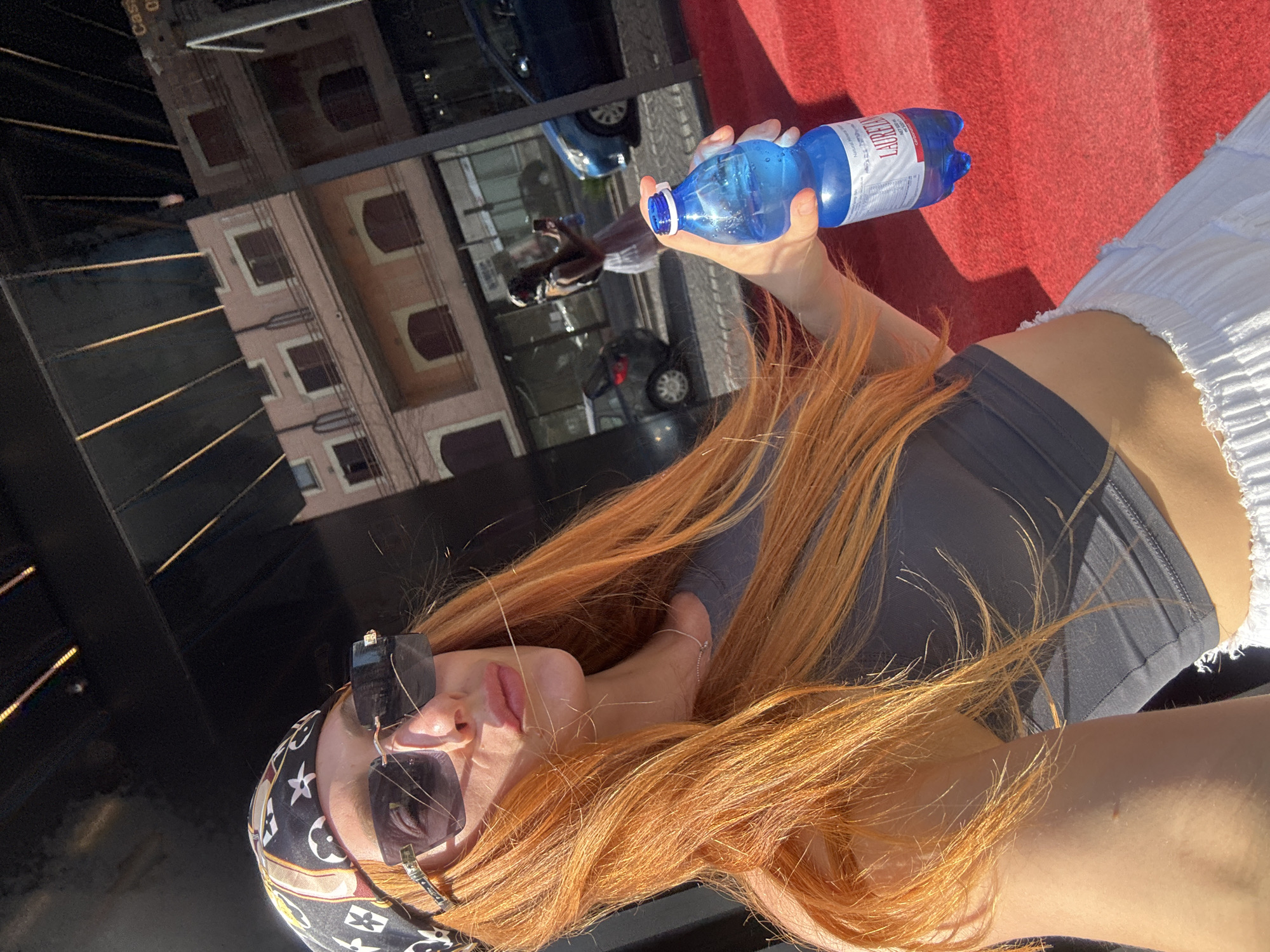 A woman with long, bright orange hair is holding a blue plastic bottle of water. She is wearing a dark grey crop top, white textured shorts, sunglasses, and a patterned bandana. She is standing on a red carpeted surface. In the background, a building with pink walls and dark red shutters is visible, along with cars parked on a street. The scene appears to be outdoors, possibly in front of a venue or establishment, with reflections in a glass surface. The context suggests this is an outdoor location in Budva, Montenegro, given the presence of a building and street.