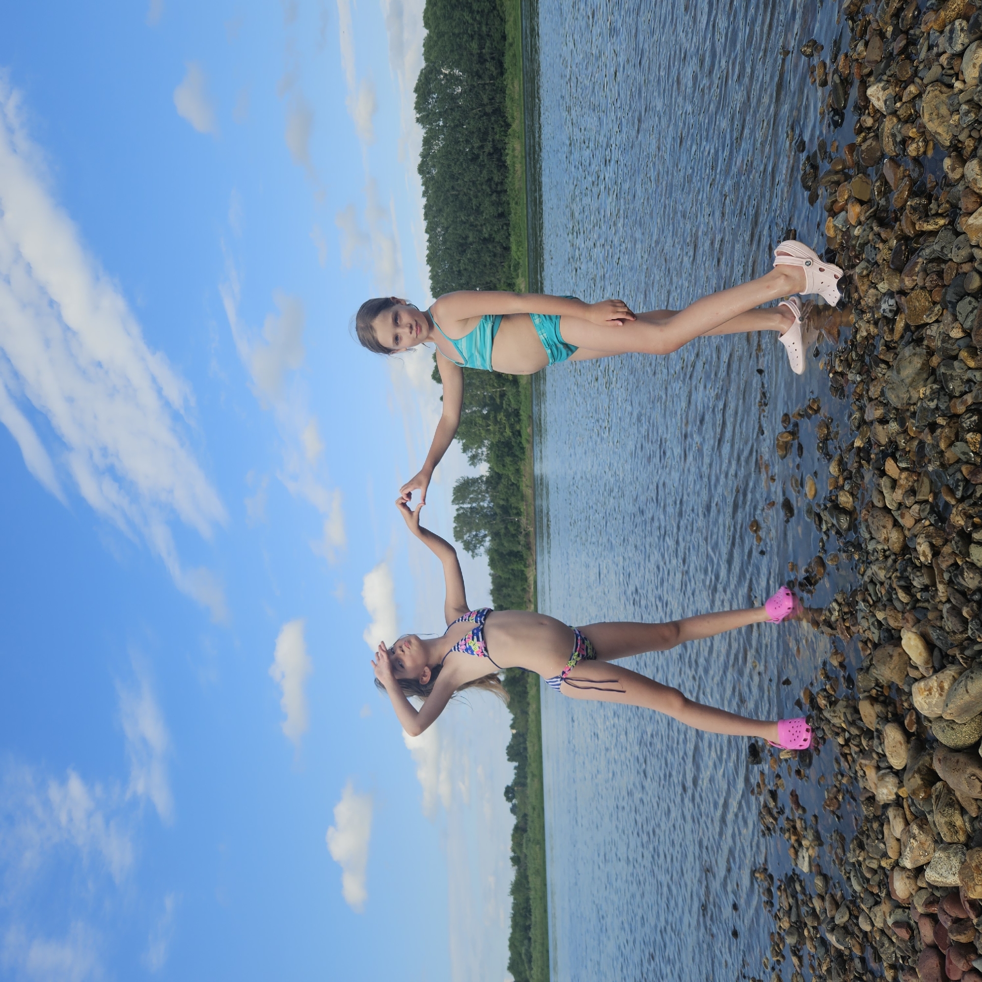 Two young girls in swimsuits are by the water's edge on a sunny day, with a clear blue sky and white clouds. The girl on top is wearing a teal and white striped bikini, while the girl below is in a multicolored floral bikini with blue stripes. The girl on top is standing on a rocky shore, wearing pink Crocs, and reaching out to the girl below. The girl below is in the water, also wearing pink Crocs, and appears to be making a heart shape with her hands. The water is calm, and a line of green trees is visible in the distance across the water. The scene suggests a leisurely summer day at a lake or river, possibly in Elykaevo, Russia, given the location context. The overall mood is playful and joyful, indicated by the girls' pose and expressions.