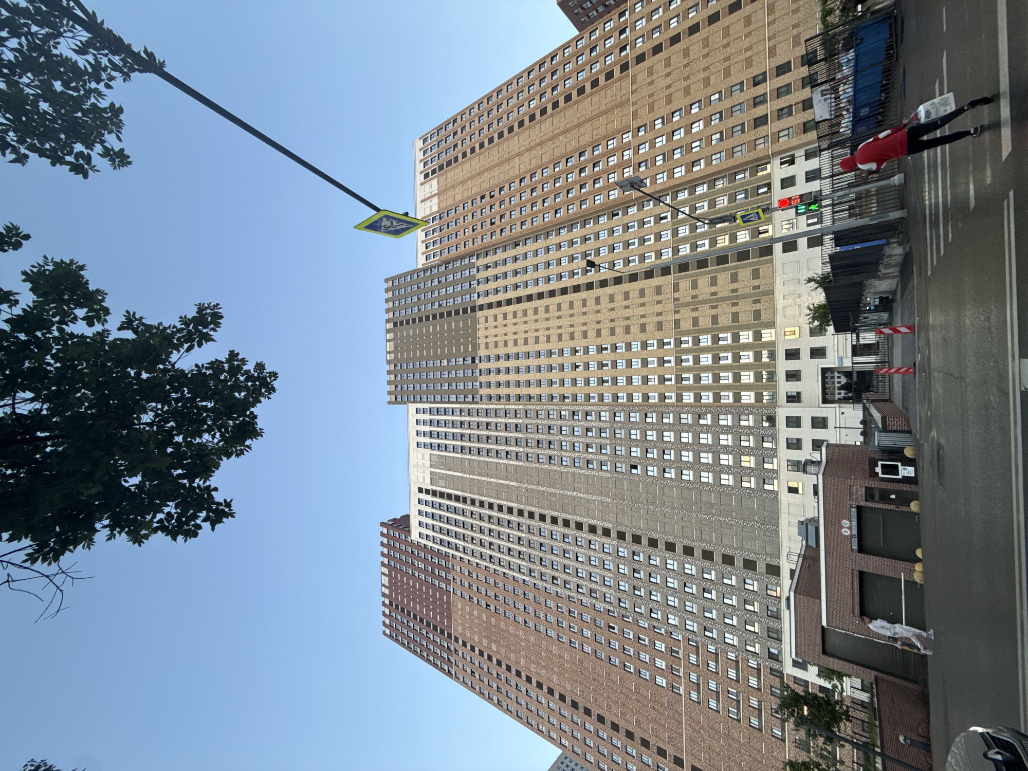 The image depicts a wide-angle view of a very tall, modern residential building complex in Moscow, Russia, under a clear blue sky. The building, characterized by its brick facade in varying shades of brown and beige, features numerous windows and is seen from a low angle, emphasizing its immense scale. In the foreground, a street intersection is visible with a pedestrian crossing and traffic lights. A person in a red hooded jacket is walking across the street, carrying a shopping bag. Another person, dressed in white overalls, is walking on the sidewalk in front of a low brick structure adjacent to the main building. Streetlights, traffic signs, and some greenery are also present. The overall impression is a contemporary urban landscape with a focus on the imposing architecture.