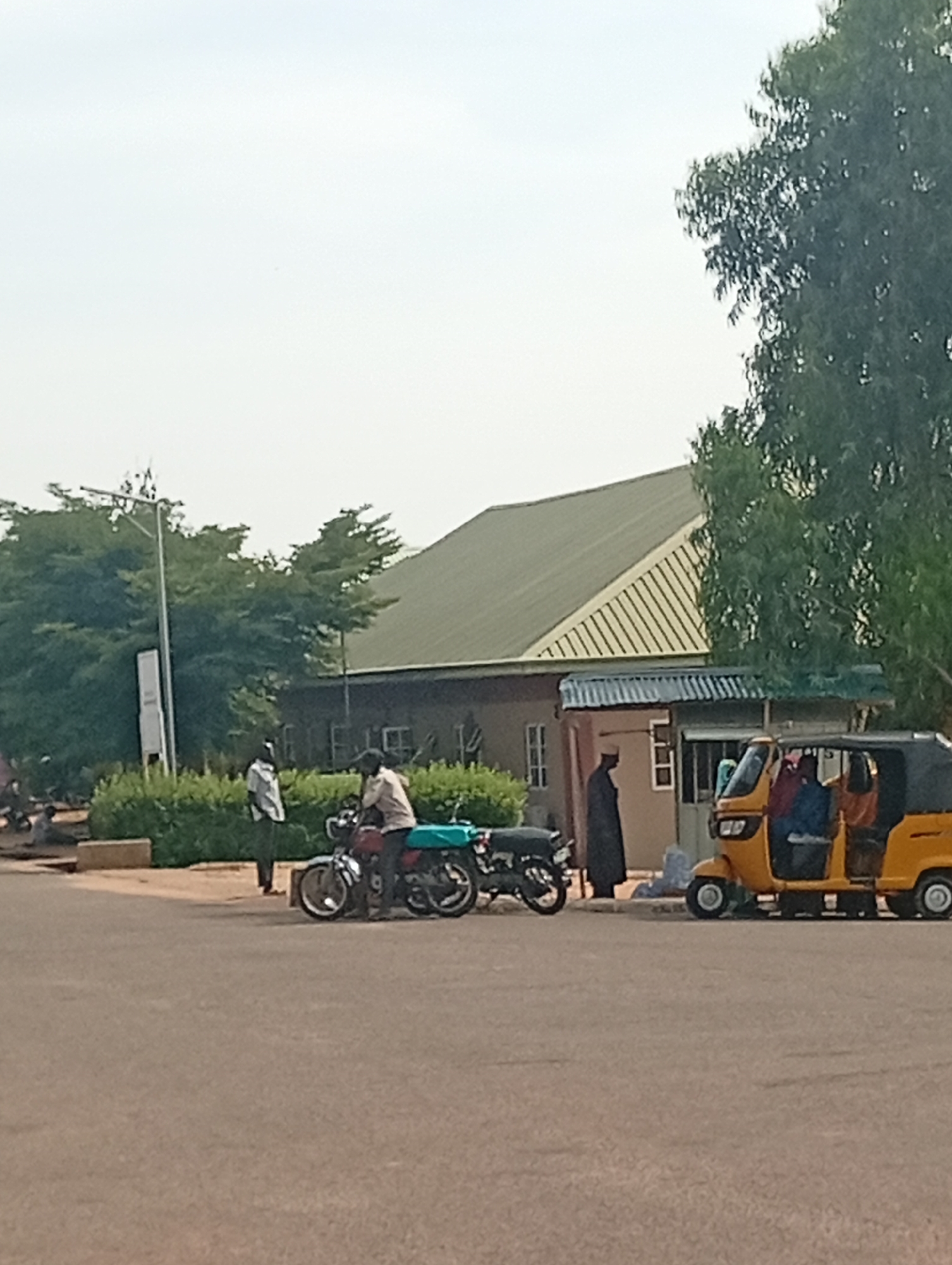 In Gombe, Nigeria, a street scene unfolds. On the right, a yellow three-wheeled auto-rickshaw, also known as a Keke Napep, is parked. Inside, two individuals are visible, one wearing a blue top and another with an orange top. A person in a dark robe stands to the left of the Keke Napep, near a small building with a corrugated metal roof.
To the left of the Keke Napep, a motorcycle is parked, with a person sitting on it wearing a light-colored shirt and dark pants. Another person, wearing a white shirt and dark pants, stands beside the motorcycle. Behind these individuals, a building with a green, sloped roof and cream-colored walls is situated. Trees and lush green bushes are visible in the background, framing the scene. Further to the left, another motorcycle is parked, and several other individuals are seated in the distance. A tall pole with what appears to be lighting fixtures is also present in the background. The ground is paved asphalt.