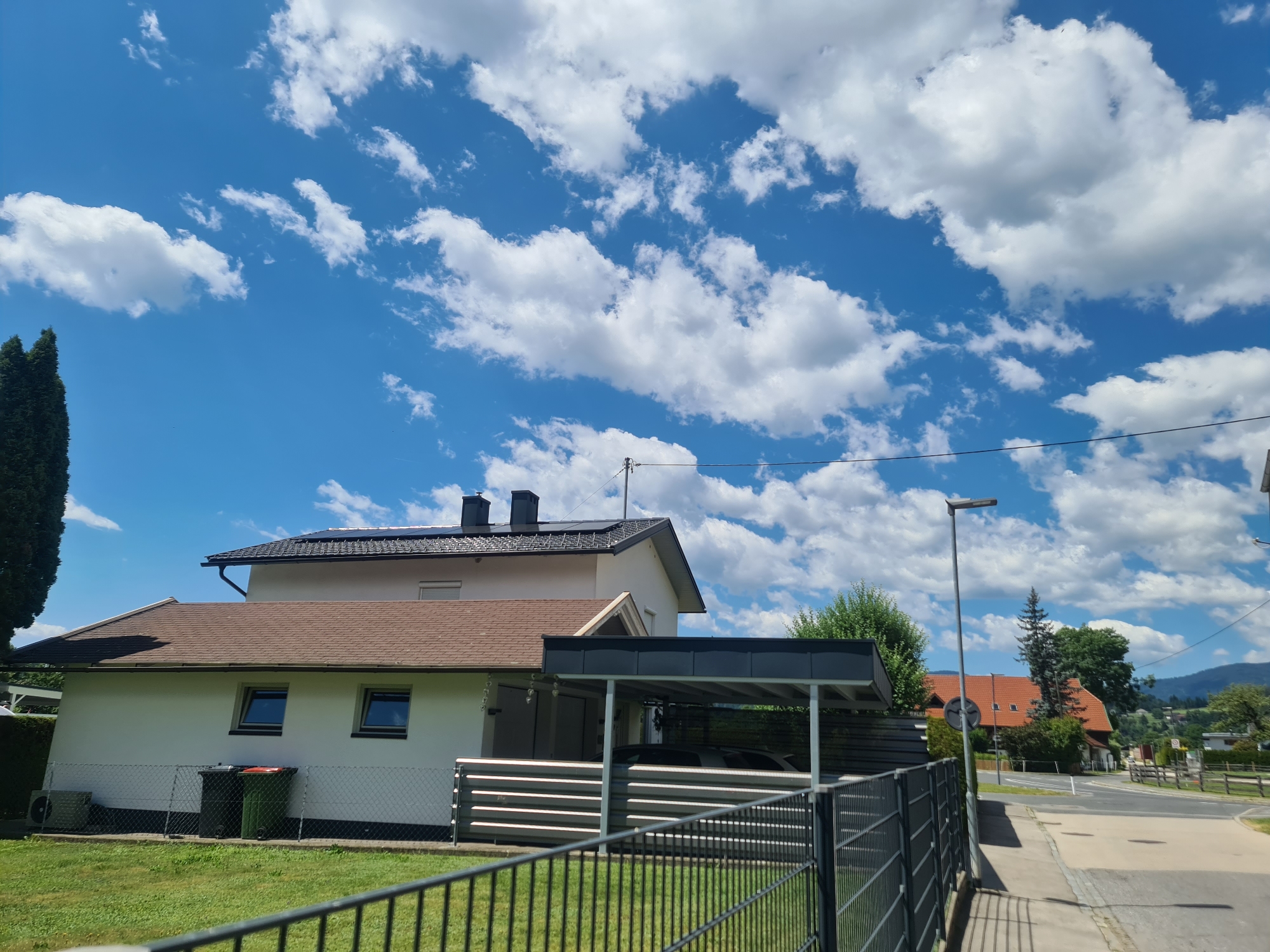The image depicts a residential area in Feldkirchen in Kärnten, Austria, under a bright blue sky filled with scattered white clouds. In the foreground, a dark metal fence runs horizontally across the bottom of the frame. To the left, a well-maintained green lawn is visible, with two large garbage bins (one dark green, one orange-lidded) situated near a white-walled house. This house has a brown-tiled roof and two small, dark-framed windows on its sloped upper wall. An air conditioning unit is visible on the ground to the left of the house.
Adjacent to this house is a covered carport with a dark grey roof and horizontal metallic slatted panels along its side. A dark-colored vehicle is partially visible under the carport. Further back, another house with a prominent orange-tiled roof and dark chimneys is visible. This house has several windows. A tall, slender evergreen tree stands to the left of this orange-roofed house.
To the right of the scene, a paved sidewalk runs alongside a road. A tall, grey street lamp stands on the sidewalk. Beyond the road, more houses and trees are visible, with a hilly, wooded landscape in the background. Power lines are strung across the upper portion of the image. The overall impression is one of a quiet, suburban or semi-rural setting on a clear day. No people are visible in the image.