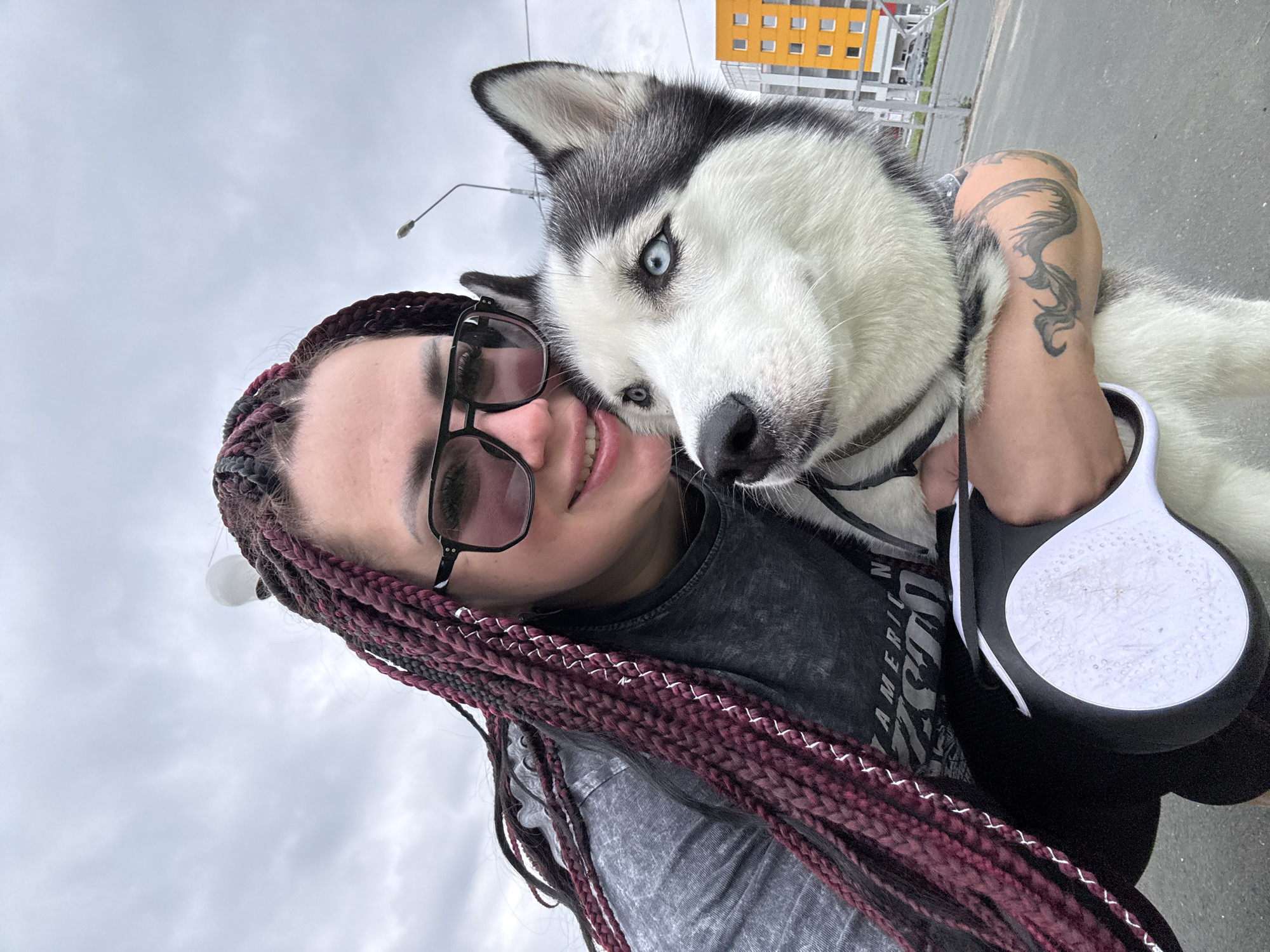 A woman with burgundy box braids and sunglasses poses for a selfie with a husky dog. The woman is wearing a gray t-shirt. The dog has blue eyes and black and white fur. Both are looking at the camera. In the background, there is an orange and yellow building and a street lamp. The scene appears to be in a city, possibly Tolyatti, Russia, given the architecture.
