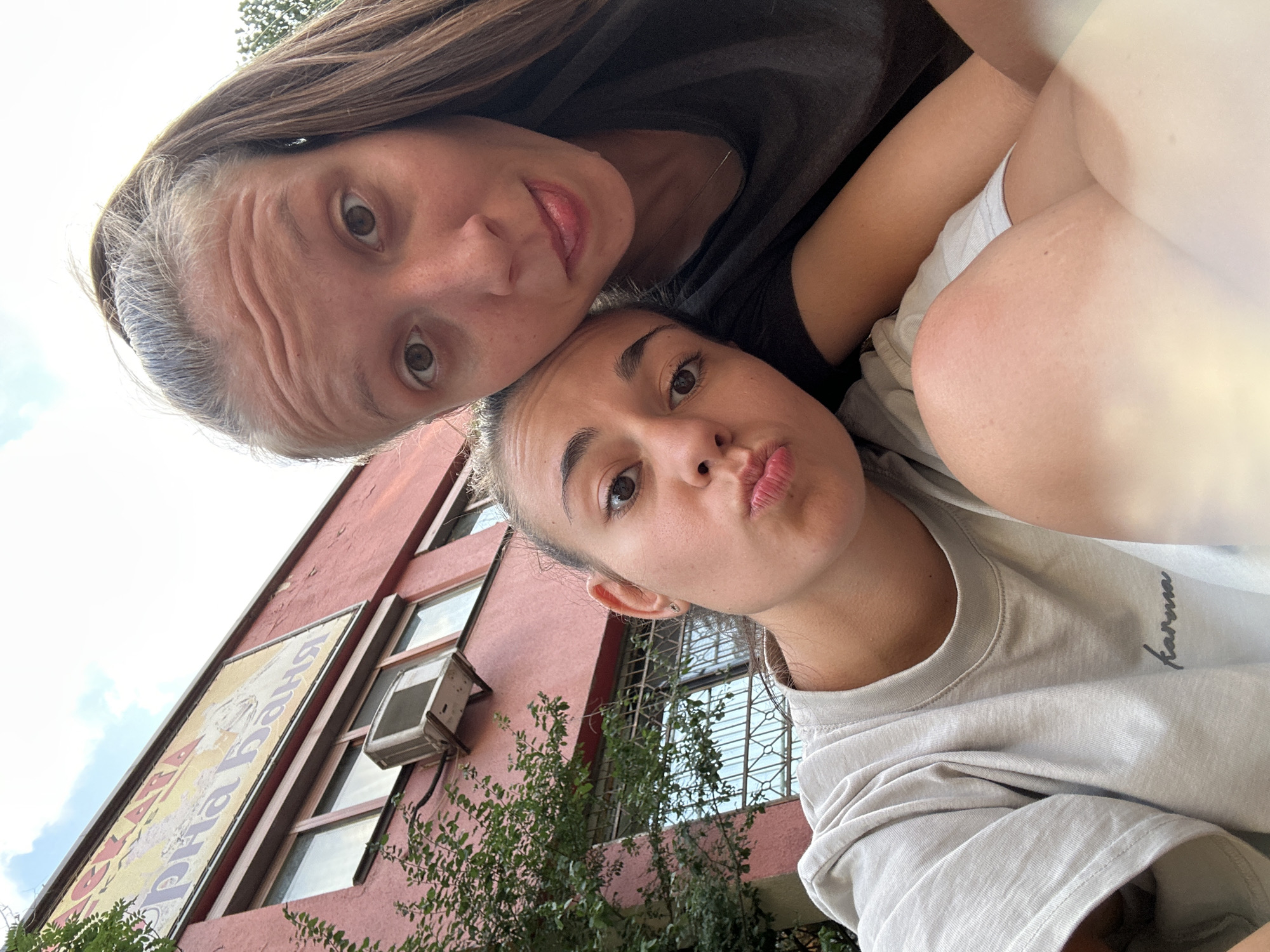 This is a close-up, low-angle selfie taken outdoors with a slightly blurred background. Two young women are the primary subjects. The woman in the foreground is on the left, facing forward with her head tilted and her lips pursed. She is wearing a light grey t-shirt with the word "karma" printed on it in cursive. Her eyes are open and looking towards the camera.
The woman behind her is on the right, leaning in and resting her head near the first woman's. She is looking directly at the camera with a slightly surprised expression, her tongue sticking out playfully. She has long brown hair tied in a ponytail and is wearing a dark t-shirt.
In the background, a building with a pink facade is visible. Above the windows, a large yellow sign with purple text is mounted. The text appears to be in Cyrillic script and reads "ЛЕКАТА ДАНЯ". An air conditioning unit is mounted on the building below the sign. Green foliage is visible on the left side of the frame, obscuring part of the building. The sky above is partly cloudy. The overall impression is a casual, candid moment captured between friends. The setting appears to be an urban environment, possibly a street or courtyard in Sofia, Bulgaria, given the Cyrillic signage.