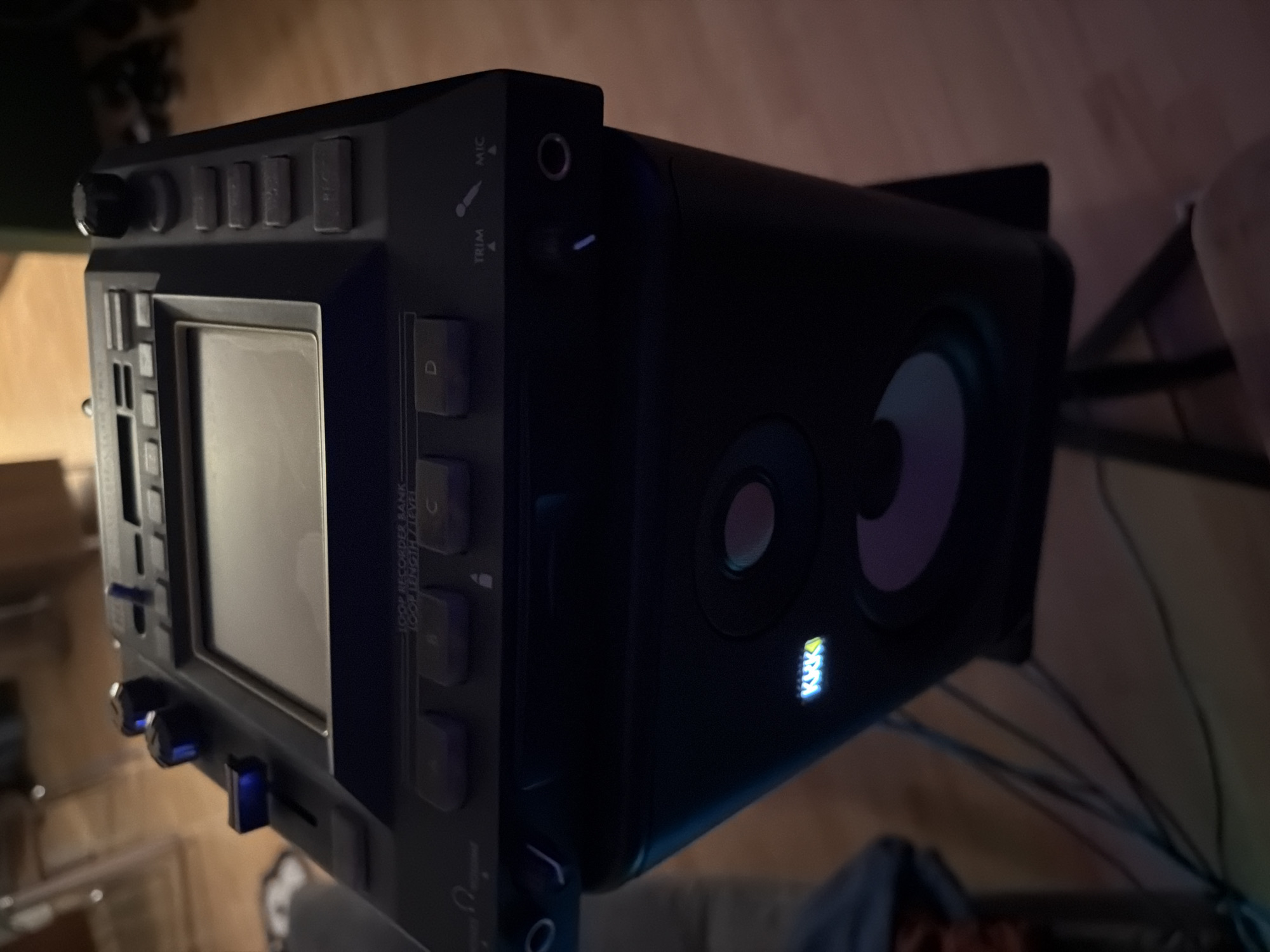 The image displays an angled, low-light shot of electronic music production equipment, likely situated in an indoor setting such as a home studio. The primary subject on the left is a dark-colored electronic device, possibly a sampler, looper, or control surface. It features a prominent central square screen or display, surrounded by numerous buttons and knobs.
Visible text on this device includes "MIC" and "TRIM" adjacent to an input jack. Other labels indicate functions such as "LOOP RECORDER BANK," "LOOP LENGTH," and "LEVEL." Four notable buttons are clearly marked "A," "B," "C," and "D." Several knobs and buttons on the device emit a distinctive blue light, highlighting them in the dim environment.
To the right, a black studio monitor speaker is visible. It features two circular speaker cones, commonly a woofer and a tweeter, and an illuminated logo on its lower front that clearly spells out "KRK." This identifies the brand of the speaker.
The setting appears to be dimly lit, suggesting evening or nighttime. A wooden surface is visible beneath the equipment, and several cables are seen trailing in the background. No people are present, and no specific activity is captured, but the arrangement of the devices strongly implies a setup for music creation, mixing, or playback. The image is captured with a slight rotational tilt, providing a dynamic perspective of the gear.
