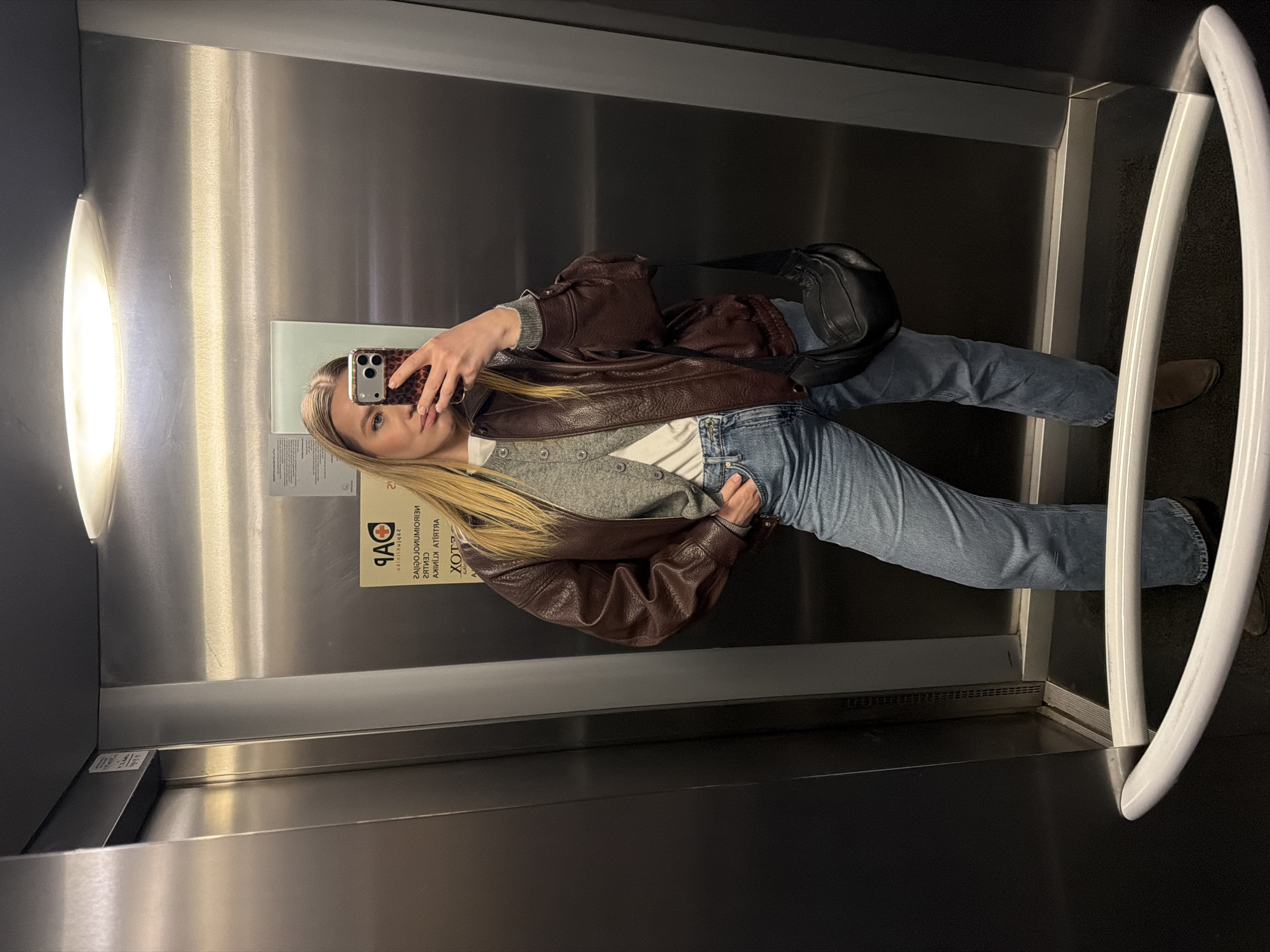 A young blonde woman in blue jeans, a white t-shirt, a gray button-up cardigan, and a brown leather jacket stands inside an elevator and takes a mirror selfie with her mobile phone, the case of which has a leopard print pattern. She is wearing a black cross-body bag. The elevator is lit by a long, vertical fluorescent light fixture and has brushed stainless steel walls. A handrail made of white tubing is visible to her right. A sticker with "QAP" and some Cyrillic text is on the wall next to her. The city and country can only be inferred from the Cyrillic text on the sticker, which is common in Riga, Latvia.