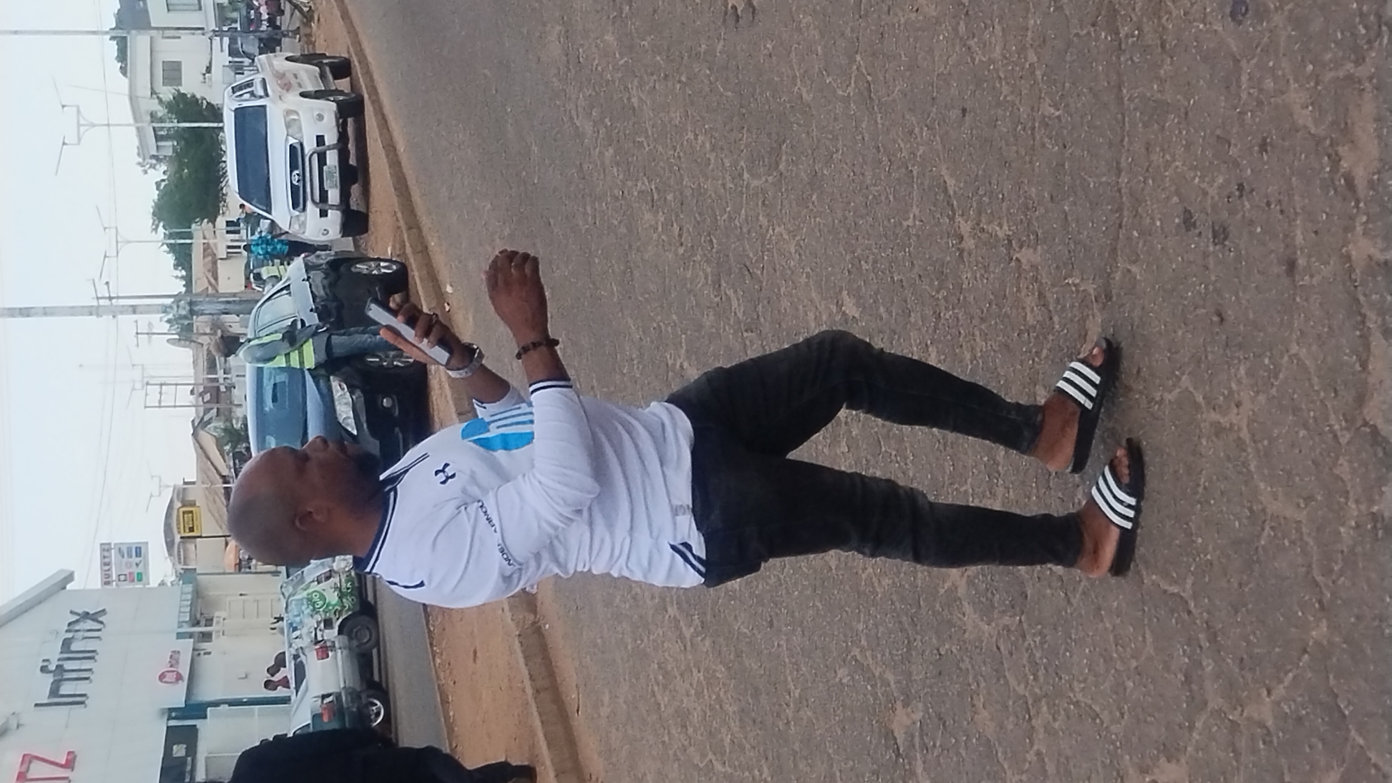 A man with a shaved head and a beard walks on a paved road in Jos, Nigeria. He is wearing a white long-sleeved shirt with a blue logo and black jeans. His feet are clad in black Adidas slide sandals with white stripes. The man is holding a smartphone in his right hand, looking down at it as he walks. In the background, a white pickup truck is parked on the side of the road. Behind the pickup truck, a black car is also parked. Further in the background, there are buildings and streetlights. The overall scene depicts a typical urban street in Jos.