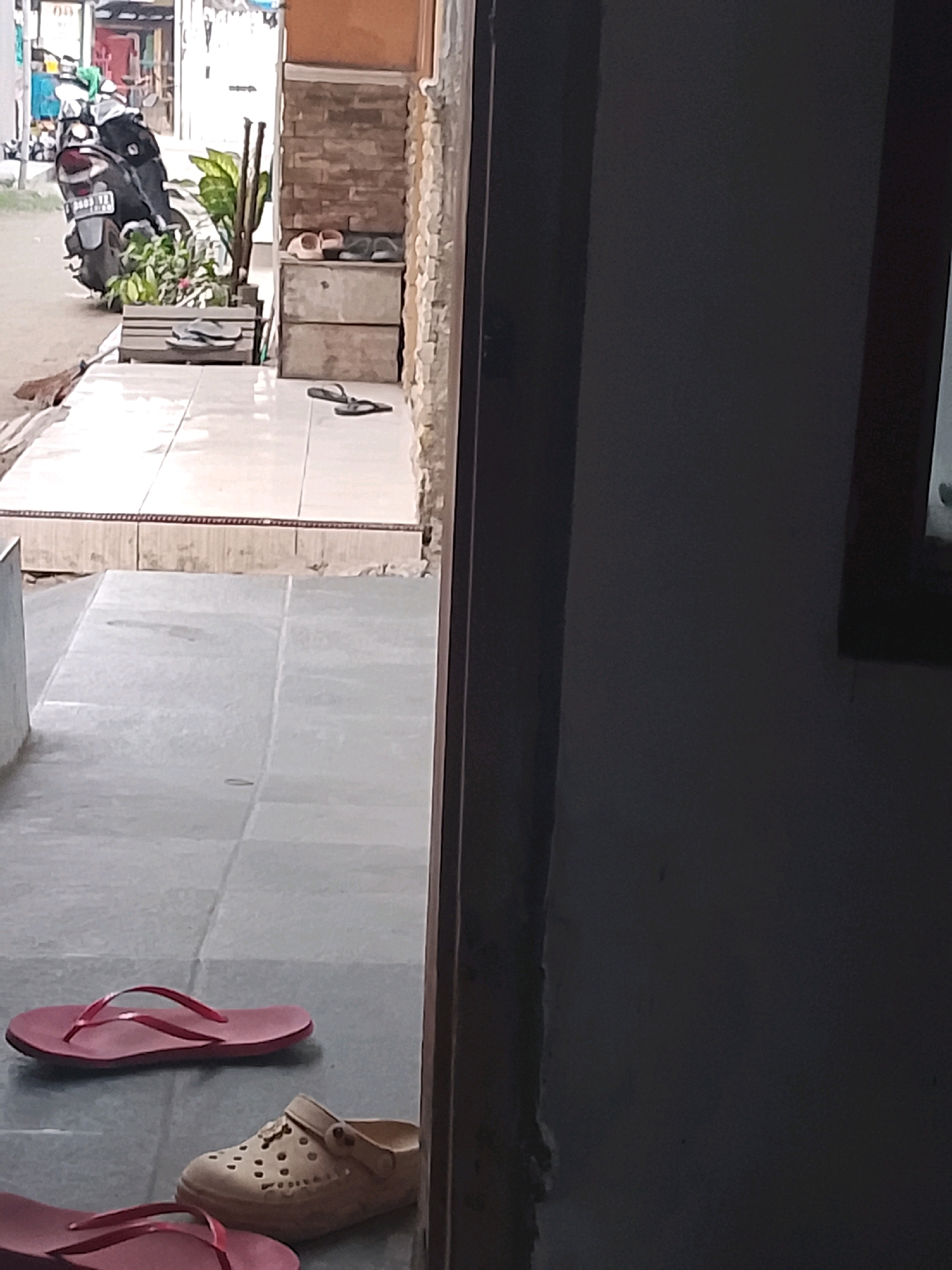 A low-angle, full-shot photograph captures an outdoor scene with a focus on footwear and a motorcycle. The foreground is dominated by a tiled floor, on which rests a pair of red flip-flops and a beige clog with decorative perforations. Slightly further back, another pair of red flip-flops is visible. To the left, a motorcycle is parked, with its rear wheel facing the viewer and its license plate displaying "B 3333 QZZ". The motorcycle is positioned near a planter box filled with greenery. Behind the planter box, a brick wall forms part of a building's facade. Two pairs of flip-flops, one pink and one dark, are neatly placed on a wooden structure. The scene appears to be in the city of Ujungrusi, Indonesia.