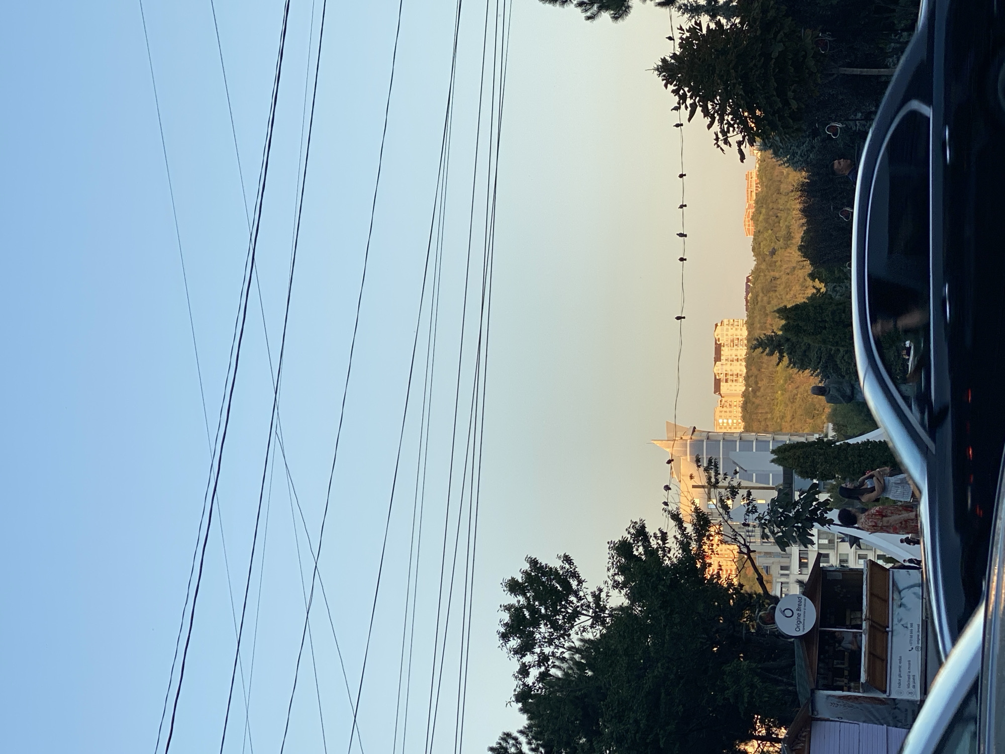The image depicts a street scene in Chișinău, Moldova, likely during sunset given the warm light. Several parallel power lines dominate the upper portion of the frame, crisscrossing against a clear, pale blue sky. A line of small birds is perched on one of these wires. In the middle ground, a modern, angular building with a white facade stands out against a backdrop of lush green hills and several multi-story apartment buildings in the distance. Some of these buildings are illuminated by the setting sun.
In the foreground, a dark-colored car is partially visible, with its reflection caught in the window. To the left of the car, a small food stall with an open counter and signage is set up. Two women are standing near the stall. Further to the right, trees with decorative red heart ornaments are visible. The overall impression is of an urban environment transitioning into a more natural, hilly landscape.