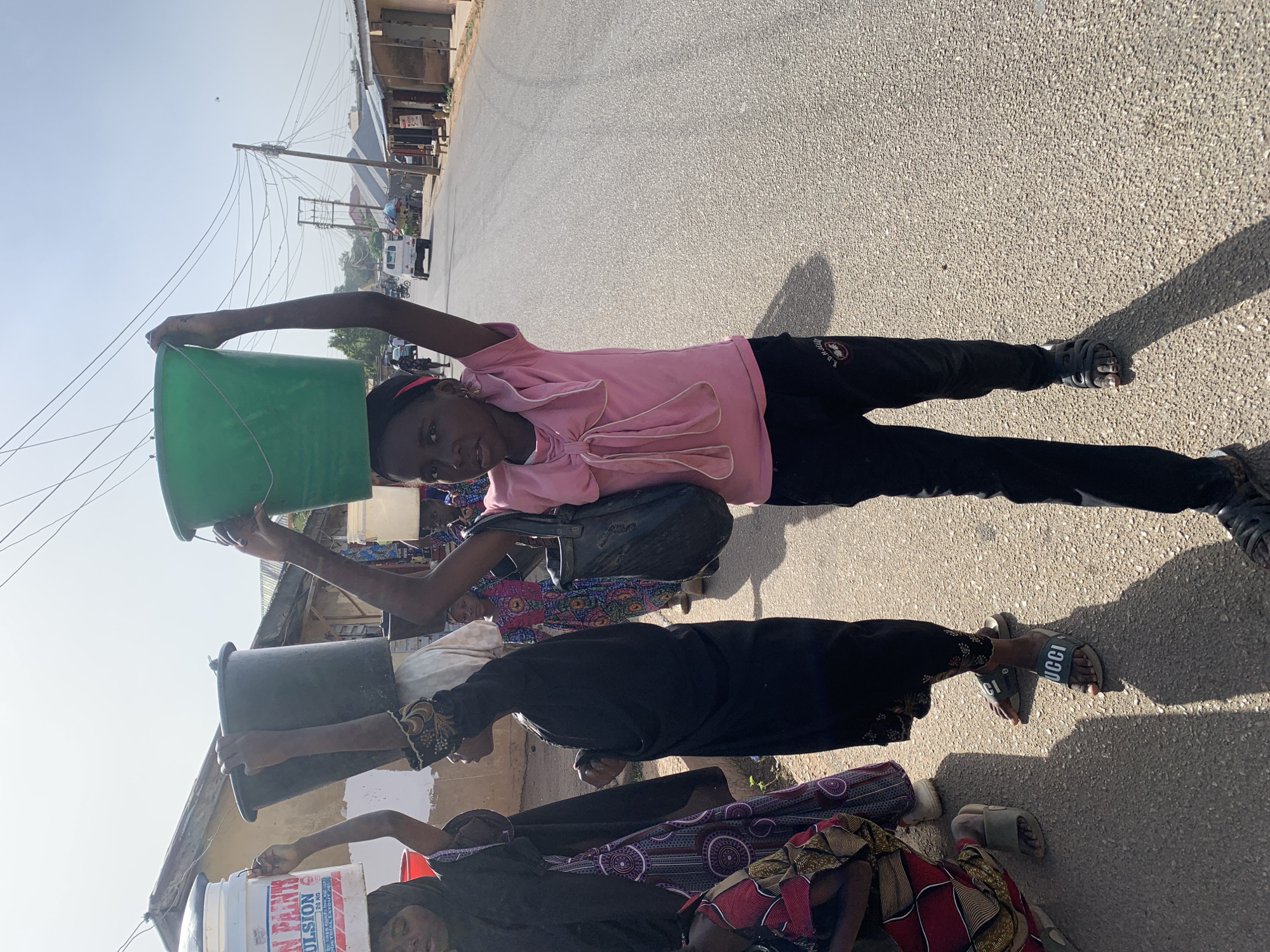 Three individuals are positioned on an asphalt road in an urban environment, likely in Jos, Nigeria. The individual closest to the camera is a child in a pink top and black pants, holding a green bucket above their head. Behind them, another person is holding a black container overhead. Further back, a third person carries a white bucket with red and blue labeling. The background includes buildings, utility poles, and a few vehicles on the street. The sky is clear and bright, indicating daytime.