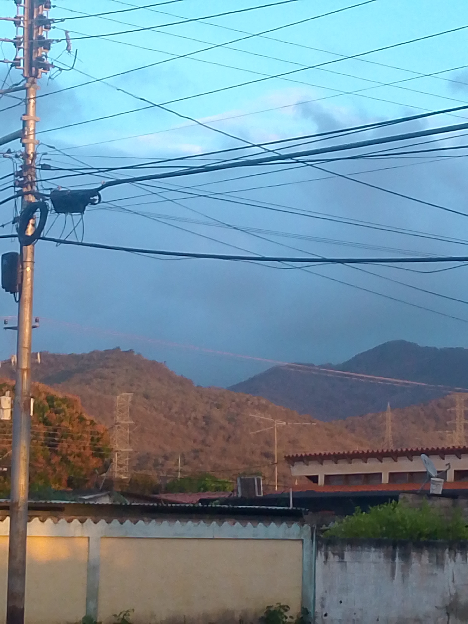 The image shows a street scene in Juan Griego, Venezuela, at what appears to be dusk or early evening. In the foreground is a concrete wall with a sawtooth top edge. Behind the wall are residential buildings with red tile roofs. Further back, undulating hills covered in brown vegetation dominate the middle ground. A power line pole stands prominently on the left side of the image, with numerous wires crisscrossing the sky. A secondary power tower is visible among the hills. The sky is a mix of light blue and darker gray, suggesting the presence of clouds or a fading sunset. There is no sign of human activity in the visible area.