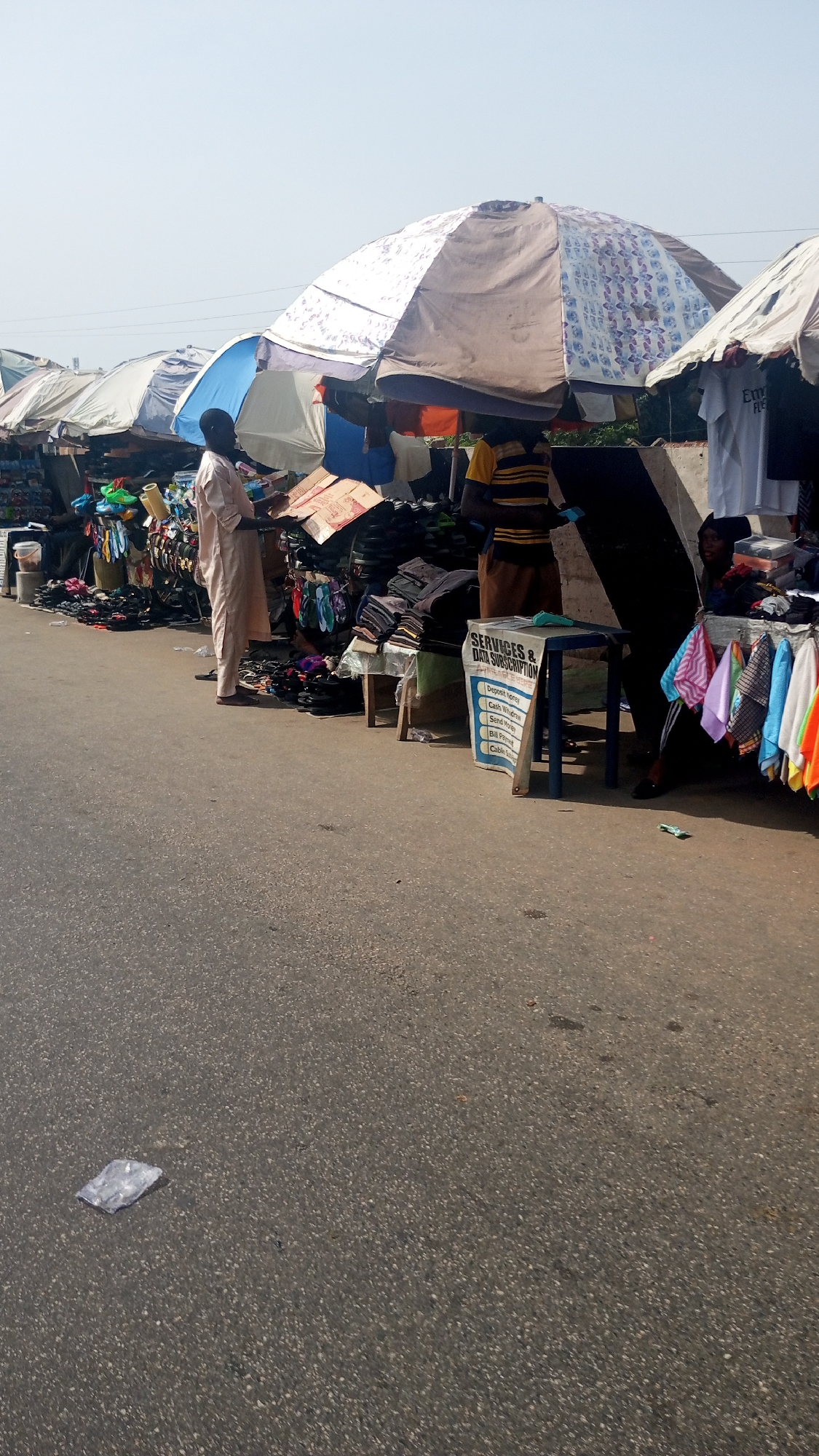 The image captures a vibrant street market scene in Lokoja, Nigeria, under bright, clear daylight. The foreground is dominated by an asphalt road, leading to a row of makeshift market stalls lined along its edge. These stalls are primarily sheltered by large, open umbrellas of varying designs, including plain ones in shades of brown and white, and patterned ones with blue and white motifs, suggesting protection from the sun.
Several people are engaged in activity. On the left, a man wearing a light-colored, long robe stands looking down at the merchandise displayed on a stall. Further right, a man in a yellow and black striped shirt and brown trousers is partially visible behind a stall, appearing to attend to customers or goods. Another individual is seated behind a stall on the far right, where colorful scarves or small cloths are neatly hung for display.
The market stalls are laden with various goods. Footwear, including sandals and slippers, is arranged in piles and on simple racks. Folded clothes and other garments are also visible, some in cardboard boxes. A prominent blue and white sign, placed on a small wooden table, reads "SERVICES & DATA SUBSCRIPTION." Below this, a list of services is visible: "Deposit money," "Cash Withdraw," "Send Money," "Bill Paying," and "Cable Subscription," indicating a mobile money or agency banking service. A piece of white plastic litter lies on the asphalt in the lower left corner of the frame. The scene conveys a bustling, everyday commercial environment.