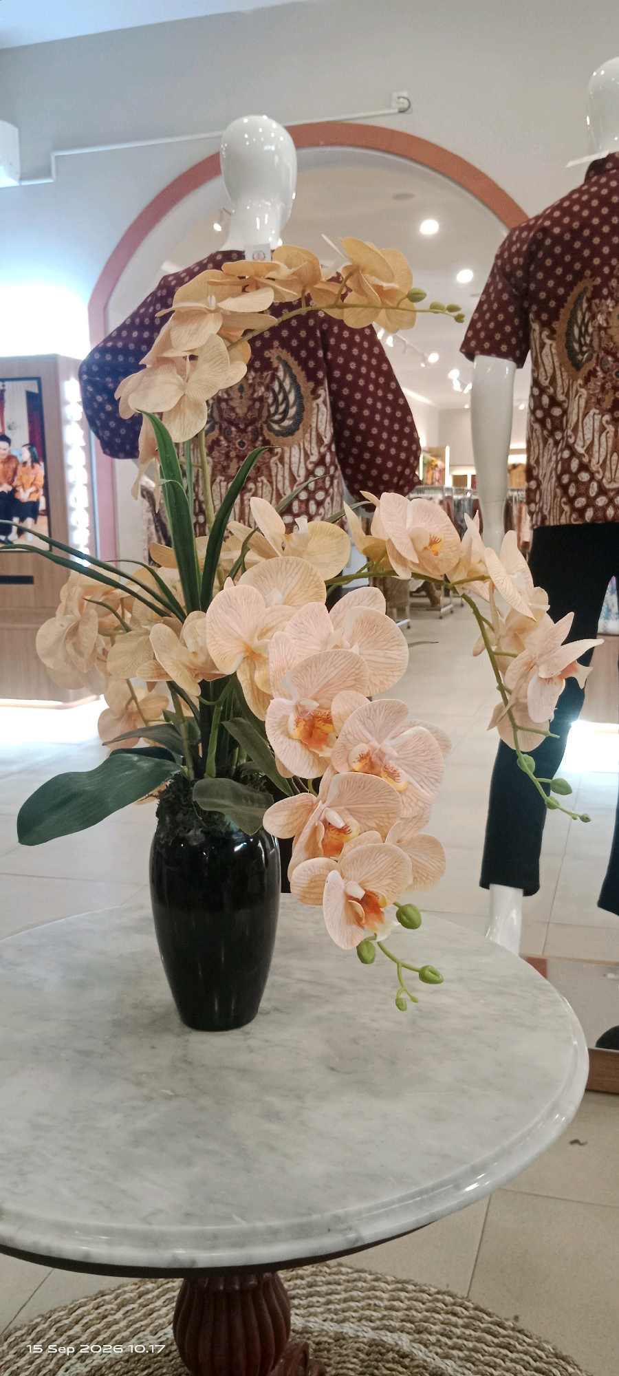 In a retail setting, a dark green vase holds a bouquet of artificial light peach orchids. The vase rests on a round marble-topped table with a decorative wooden base. In the background, two mannequins are displayed, each wearing a maroon batik shirt with a pattern of small dots and a more elaborate batik design across the chest and shoulders. The mannequins are part of a retail display, with clothing racks visible in the distance. The flooring appears to be tiled. The timestamp in the lower left corner indicates the image was captured on September 15, 2026, at 10:17. The location is identified as Surakarta, Indonesia.
