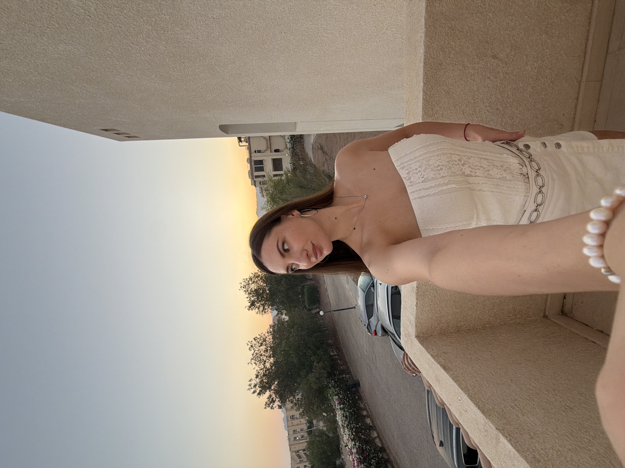 A young woman with long brown hair is taking a selfie on a balcony in Sharm El Sheikh, Egypt. She is wearing a white strapless top and a white skirt with a silver chain belt. The background shows a street with parked cars, trees, and buildings. The sky is a pale blue with hints of orange and yellow, indicating it is either sunrise or sunset.