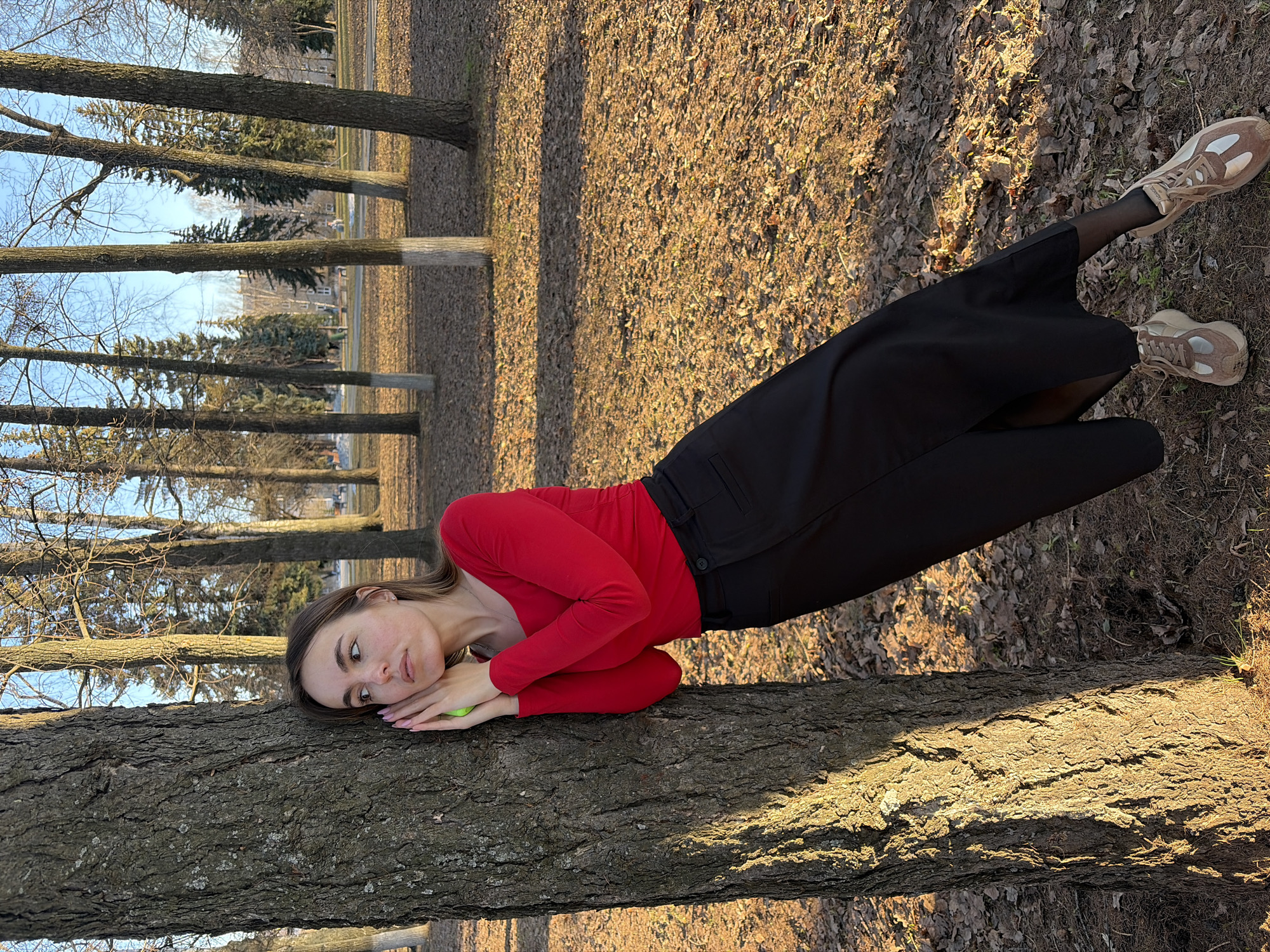 A young woman is reclining on a fallen tree trunk in a park. She is wearing a long-sleeved red top, black pants, sheer black tights, and light brown sneakers. Her head is resting on her folded hands, and she is holding a yellow-green object in her hands. The park is visible in the background, featuring other trees and ground covered in leaves. The location appears to be Izhevsk, Russia.
