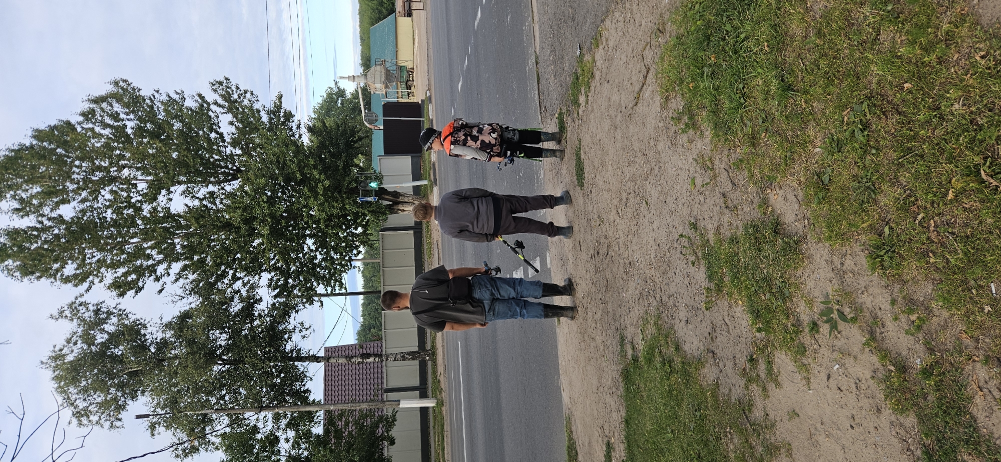 The image shows three people, likely two adults and one child, standing on a dirt and grass area next to a road. They are all facing away from the camera and appear to be holding fishing rods. The person on the left is wearing a dark t-shirt, blue jeans, and tall black boots. The middle person is wearing a grey sweatshirt and dark pants. The person on the right, appearing to be a child, is wearing a camouflage jacket with an orange vest and a black hat. A traffic light with a green signal is visible in the background, suggesting the location is an intersection. The environment includes trees, a fence, a building with a brown roof, and power lines. The overall scene suggests a casual outdoor activity, possibly fishing. The image does not contain any specific landmarks or identifying features that would definitively place it in Vitsebsk, Belarus.