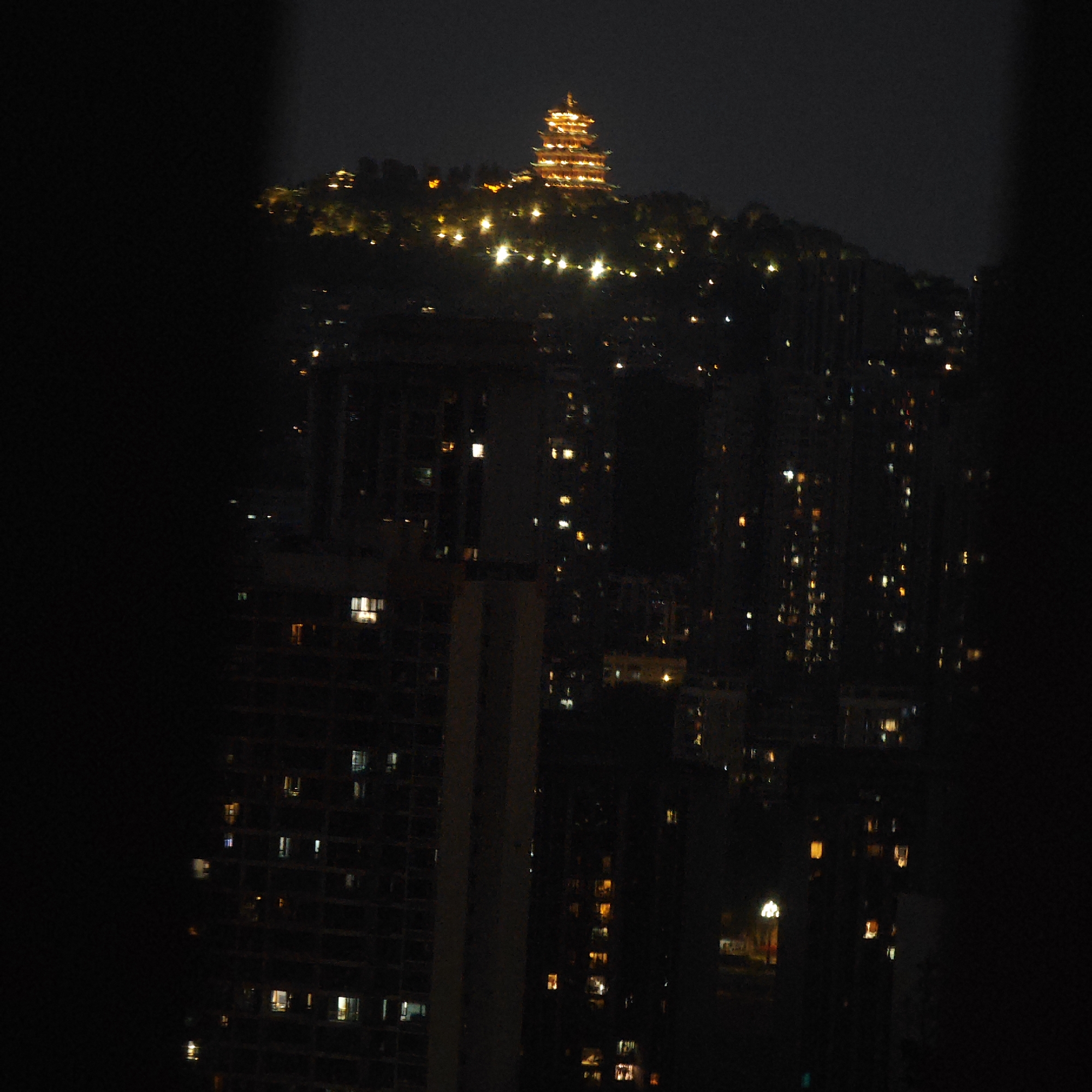 A nighttime shot, heavily underexposed, focuses on a pagoda atop a hill in the Yuzhong District of China. The pagoda and the hillside are illuminated with orange lights, contrasting with the darker surroundings. Below, a cluster of high-rise buildings is visible, punctuated by numerous lit windows. The composition is partially obscured by dark, out-of-focus elements in the foreground, suggesting the scene is viewed from an elevated position. The overall image conveys an urban landscape at night.