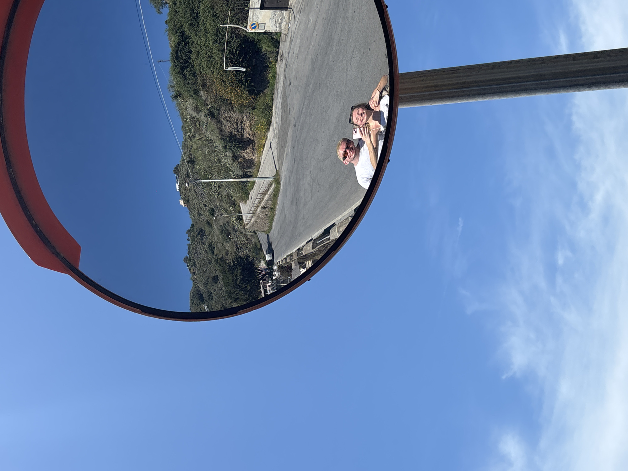 Captured from a tilted perspective against a clear blue sky with scattered clouds, the image centers on a convex traffic mirror mounted on a metal pole. The mirror's orange rim prominently frames its reflective surface.
The reflection shows a road bordered by a hillside covered in greenery, and a couple posing for a picture, with one holding a phone. The reflection suggests that the photograph was taken near Lipari, Italy, considering the local flora and architecture visible. The individuals are wearing light-colored casual clothing and sunglasses, looking directly at the camera phone. The mirror is positioned to provide visibility of the bend in the road and the adjacent hillside.