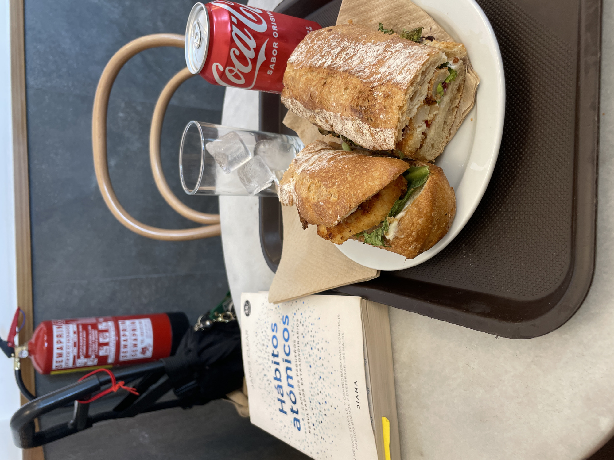 The image shows a brown plastic tray on a light-colored tabletop. The tray holds a white plate with two halves of a sandwich, a can of Coca-Cola, and a glass of water with ice. Next to the tray is a book titled "Hábitos atómicos". A red fire extinguisher is partially visible in the lower left corner of the image, along with a black umbrella. The image likely depicts a meal setting in a casual environment. There is nothing to suggest that this photo was taken in Barcelona, Spain.