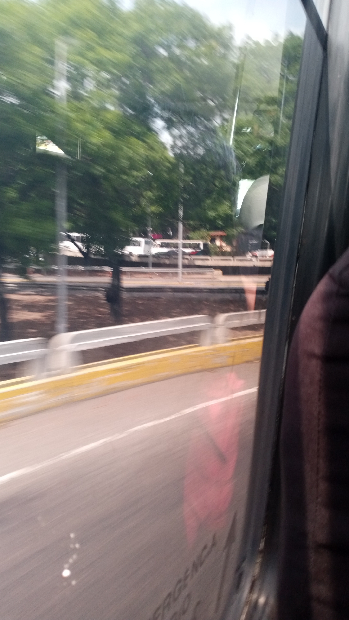 The image shows a view from inside a moving bus, looking out the window. The outside scene includes a road, a barrier with yellow paint, and trees. There are also some vehicles visible in the distance, including a white truck. The foreground includes the bus window and part of a seat. The word "EMERGENCIA" is visible on the bus window. The location is Unknown, Unknown.