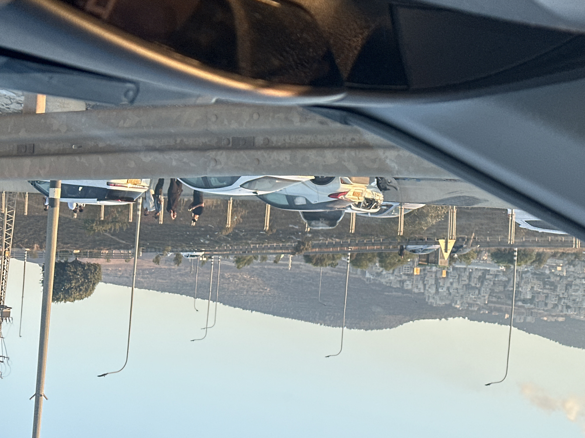 A number of vehicles, including a white SUV, are parked on the side of a road. Several people, some appearing to be men, are standing near the vehicles. The scene is set against a backdrop of a city built on a hillside, with mountains in the distance. The sky is clear and a warm light suggests it is either sunrise or sunset. The area appears to be in Turan, Israel, indicated by the landscape and architectural style of the buildings on the hillside. The presence of multiple vehicles and people suggests a possible gathering or event.