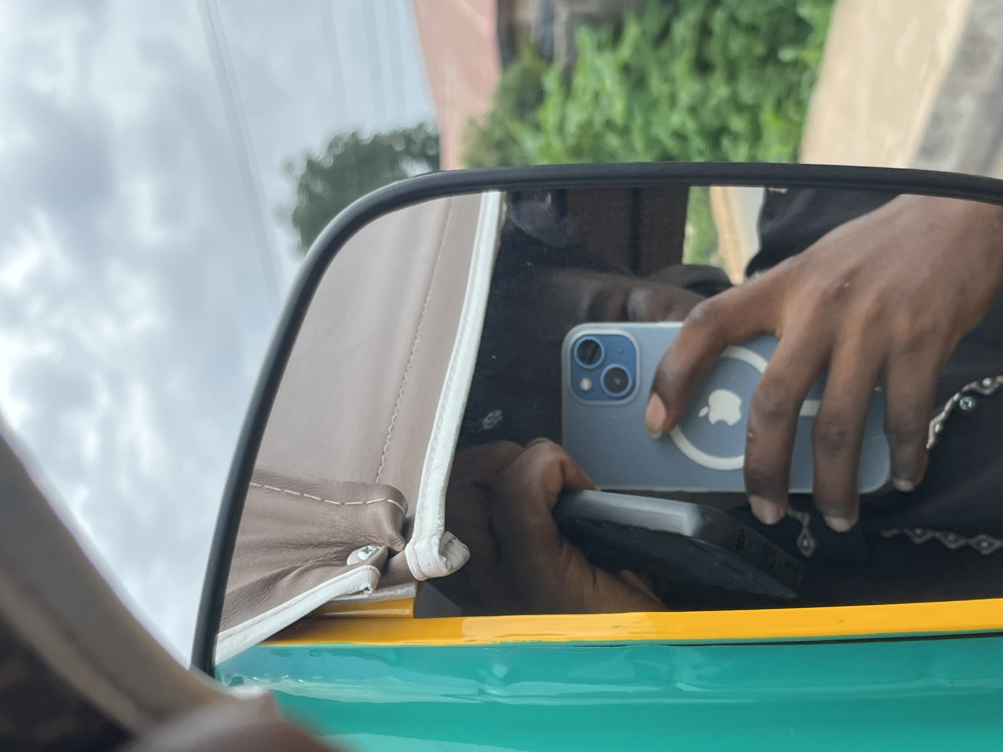 A reflection in a side-view mirror of a vehicle shows a person holding a light blue smartphone with an Apple logo. The person is wearing a dark garment. The mirror is framed by the vehicle's turquoise and yellow exterior. The background reflection shows a cloudy sky, trees, and buildings, suggesting an outdoor urban environment. The image appears to have been taken in Bukuru, Nigeria.