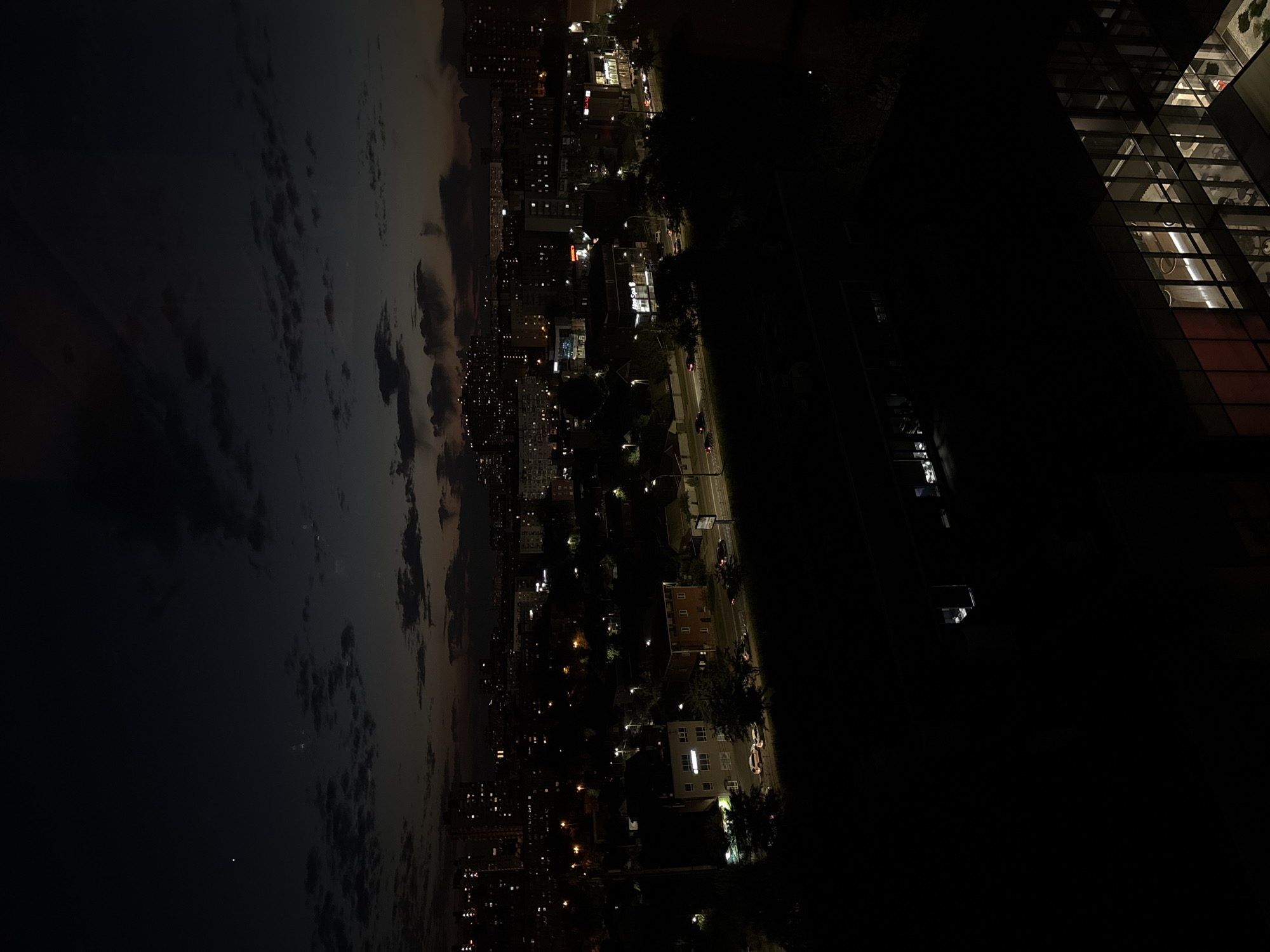 The image depicts a city at dusk or early night, with a mix of artificial lights and natural twilight. The perspective is elevated, suggesting a view from a building or a high vantage point. On the left side, a vast expanse of cloudy sky dominates, with hints of lighter hues near the horizon indicating the fading daylight. Scattered across the sky are patches of darker clouds, creating a textured appearance.
The right side of the image reveals an urban landscape. Buildings of varying heights are visible, many with illuminated windows, indicating occupancy. Streetlights line roads, casting pools of light. Some vehicles can be seen on the streets, their headlights and taillights creating streaks of light. Trees are scattered throughout the urban area, their forms silhouetted against the lights. In the foreground, a modern building with a glass facade is prominent, its interior lights creating a geometric pattern. The overall scene suggests a bustling city winding down for the night or just beginning to come alive with its evening illumination. The time of day is clearly evening or night, with the presence of artificial lighting and the fading natural light. The weather appears to be partly cloudy. The location is identified as Krasnodar, Russia. There is no visible text in the image.
