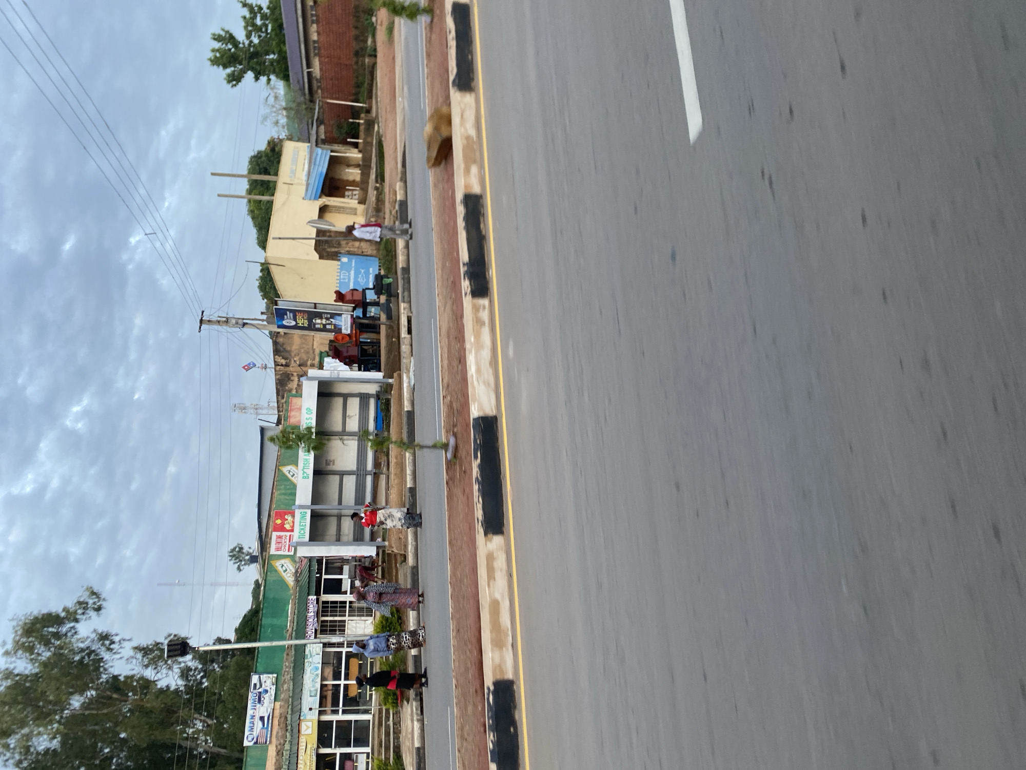 The image shows a street scene in Jos, Nigeria. In the foreground, a paved road with a yellow line and a white marking runs horizontally. A concrete curb with black and white markings separates the road from the sidewalk. On the sidewalk, several people are standing and walking. To the left, a green building with "MAN-JIMON" and "PROVISION STORE" signage is visible. To the right of the provision store, there is a bus stop shelter with a white roof and benches. A man in a white shirt and trousers stands near a large rock on the side of the road. In the background, there are more buildings, trees, and power lines under a cloudy sky. The overall scene suggests a typical day in a Nigerian urban environment.