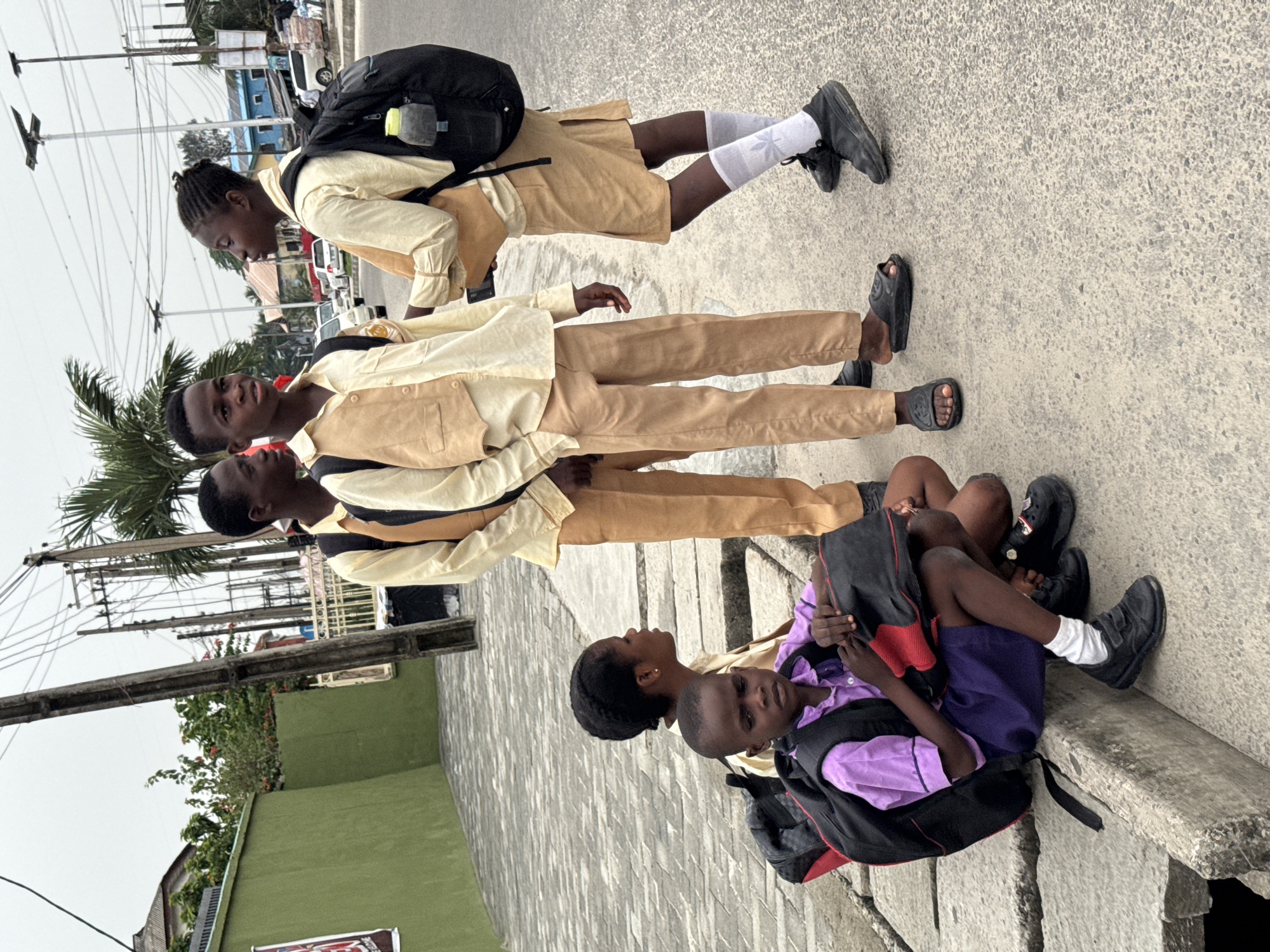 Ado, Nigeria, presents a street scene featuring a group of students. Three individuals are seen standing on a sidewalk, one wearing a uniform consisting of a light yellow shirt, a tan vest, and tan pants, complemented by black sandals, while the others are in similar attire. Additionally, a person at the end of the sidewalk is seated, also in a school uniform. One student has a backpack. They are all standing on a gray sidewalk. In the background, there are buildings, trees, and power lines visible. The sky is overcast.