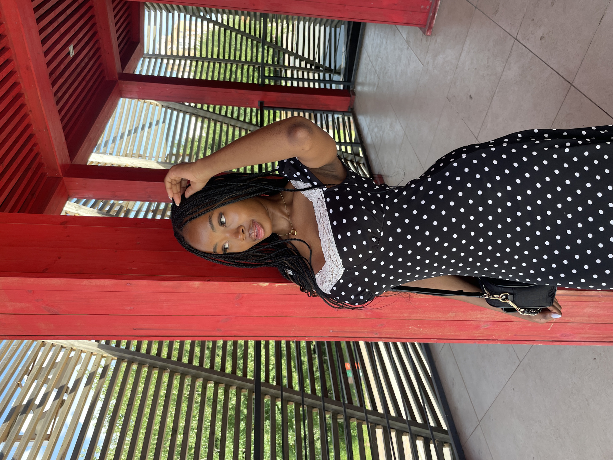 A young Black woman with braided hair stands in a covered outdoor area with red wooden beams and a slatted wooden roof. She is wearing a black and white polka dot dress with white lace trim around the neckline. A black purse with a gold chain strap is slung over her left shoulder. She is touching her hair with her right hand and looking to her left with her mouth slightly open. The background shows a light-colored tiled floor and a view of green trees and a blue sky through the slatted wooden structure. This location is the Kremlin Embankment in Kazan, Russia, specifically the "Krasnye Nitsy" (Red Threads) art object.