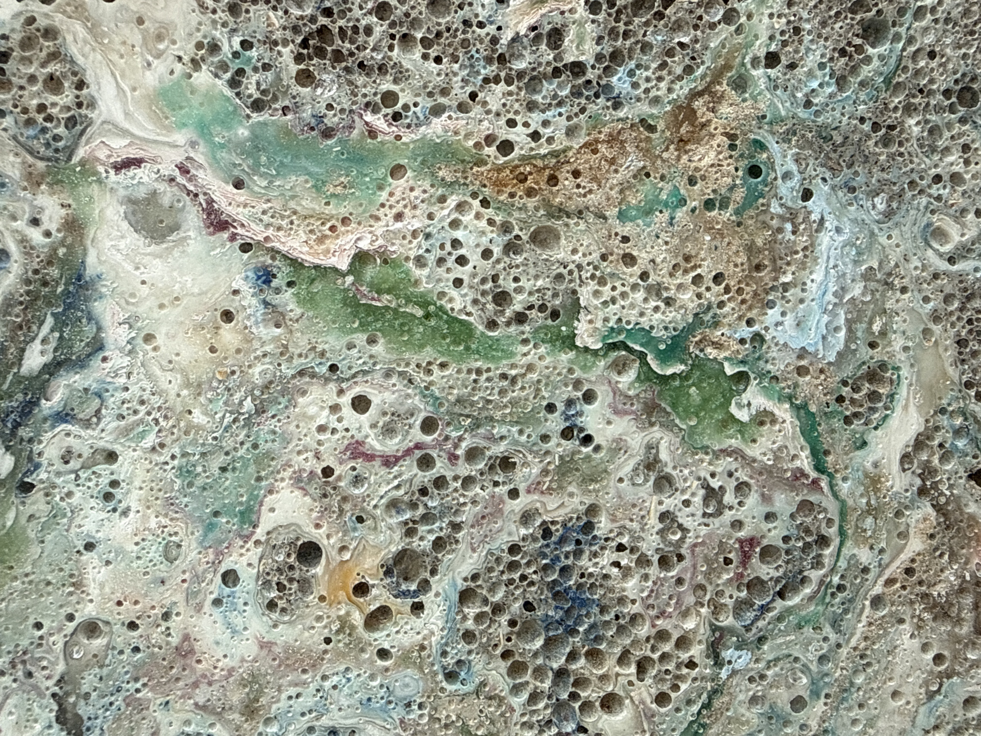 The image depicts a close-up abstract texture, likely of a painted or sculpted surface. The dominant colors are off-white, grey, and green, with hints of blue, brown, and purple. The texture is characterized by numerous small holes or air bubbles, creating a porous and uneven surface. The colors appear to be mixed and swirled, suggesting a fluid application or natural formation process. The composition lacks any identifiable human figures, clear objects, or interactions. The environment is limited to the immediate surface of the material, precluding any information about a specific location like Dubai, United Arab Emirates.
