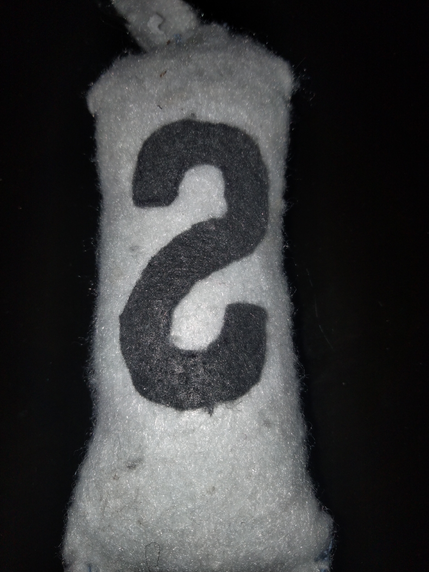 A close-up, top-down shot of a stuffed toy with the number "2" in dark gray felt sewn onto a white, fuzzy fabric body. The background is entirely black. The toy is lying on its side, and its shape suggests it may be cylindrical or elongated. The lighting is direct and focused on the toy, creating shadows and highlighting the texture of the fabric. There are no people or other objects visible in the frame. The image is taken with a macro lens, emphasizing the details of the stitching and fabric fibers. No information about the location in Payagaboh, Indonesia, can be derived from the image itself.