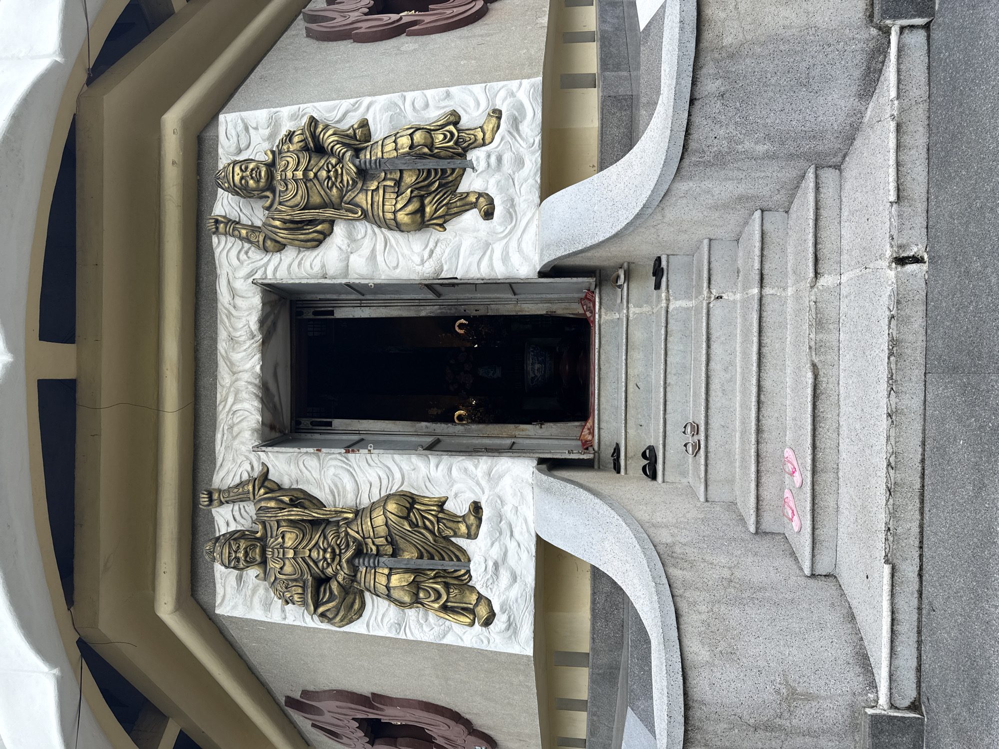 The image depicts the entrance to a Buddhist temple in Nha Trang, Vietnam. Two large, golden statues of temple guardians, each holding a sword, flank the doorway. The statues are mounted on white relief carvings depicting clouds. In front of the entrance, a set of granite stairs leads up to the doorway. Scattered on the stairs are several pairs of flip-flops, some pink, some black, and some light brown. The overall architecture suggests a traditional Southeast Asian temple design.