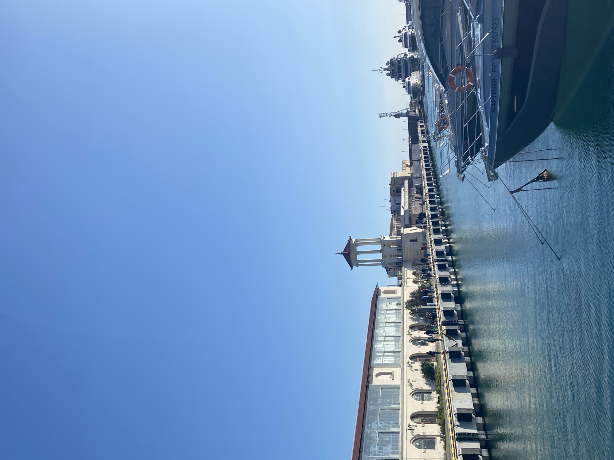 The image captures a vibrant, sunlit harbor scene, likely in Sochi, Russia, under a clear blue sky. The perspective looks along a pier, revealing various vessels and architectural structures.
A vast, unblemished light blue sky fills the upper portion of the frame. Below this, the calm, blue-green waters of the harbor gently reflect the bright sunlight.
Along the right side of the image, a concrete pier extends into the distance, lined with various boats and ships. In the foreground, the bow of a large, dark-hulled boat is prominently displayed, showing its railings and an orange life buoy. The name "МАРТИНИ ВЕ" (MARTINI VE) is clearly visible on its hull, accompanied by the registration "17 RUS23." A small, dark pennant or flag hangs near its bow. Further along the pier, several larger ships, potentially patrol boats or military vessels, are docked, identifiable by their intricate mast structures, radar domes, and communication equipment.
On the left side, running parallel to the pier, a series of buildings form the backdrop. A multi-story structure with a light-colored facade features arched windows on its lower levels and modern glass sections above. Further down the pier, a distinctive white, classical-style pavilion or rotunda, complete with columns and a reddish, peaked roof, stands out.
The scene depicts a peaceful, bright day at the port. The clear sky and calm water suggest excellent weather. No specific activities or individuals are distinctly visible, portraying a quiet moment in an otherwise active maritime setting.