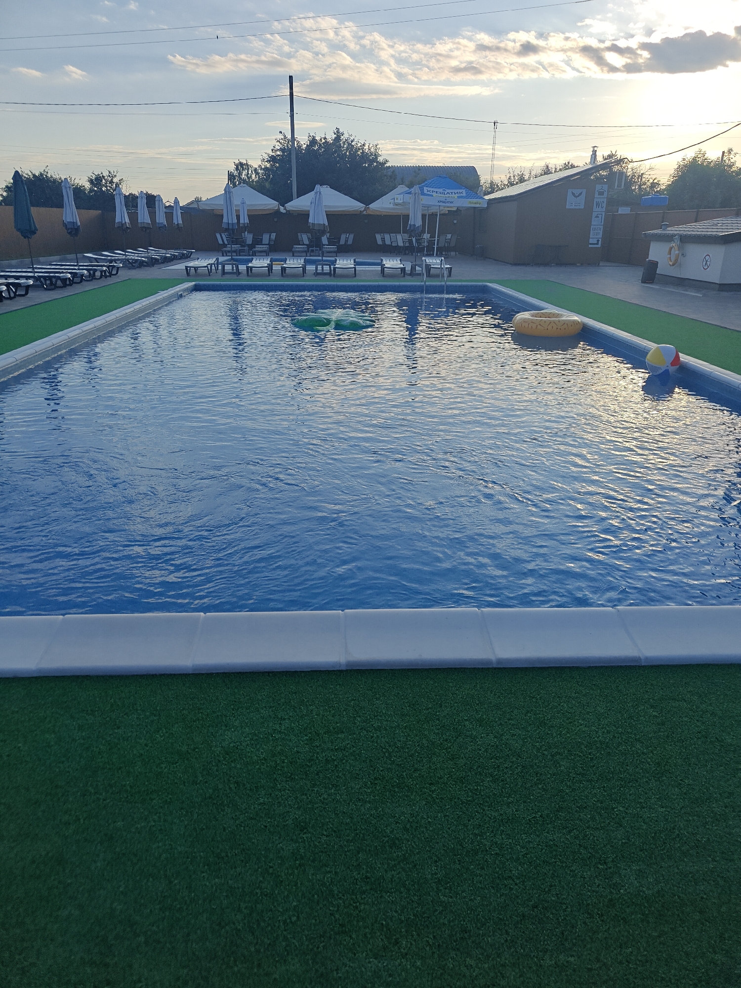 The image depicts an outdoor swimming pool area at a resort located in Shkurupiivka, Ukraine, bathed in the soft light of dusk. The main subject is a rectangular swimming pool filled with clear blue water. Floating in the pool are a yellow inflatable ring and a colorful beach ball. The pool is surrounded by a white tiled border, and beyond that, a strip of artificial green turf. In the background, rows of lounge chairs and closed umbrellas are neatly arranged on paved ground. Further back, a wooden building with a sloped roof and signs, including a "WC" symbol, stands under a partly cloudy sky. Trees and a utility pole are visible on the horizon. The overall scene suggests a peaceful and serene atmosphere, possibly after a day of activity at the resort.