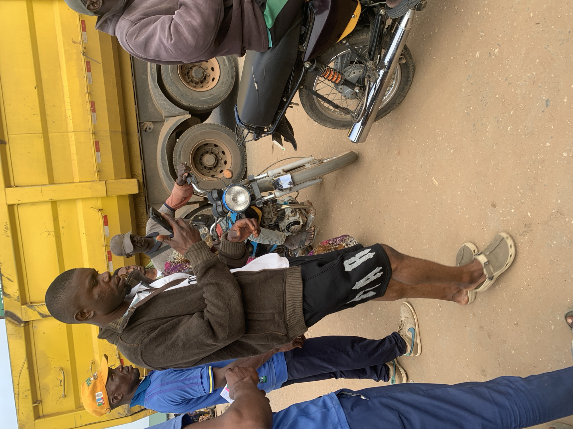 In Shagamu, Nigeria, on a dirt road, a man in a brown jacket and black shorts is looking at his phone while standing next to a yellow dump truck. He is wearing tan clogs. To his left, a man in a blue shirt and dark pants is also looking at his phone. Behind them, another man in a cap and a dark jacket is visible. A motorcycle is parked to the right of the man with the phone, and another motorcycle is parked to the left of the dump truck. The dump truck is yellow with reflective red and white stripes. The ground is sandy and dusty.