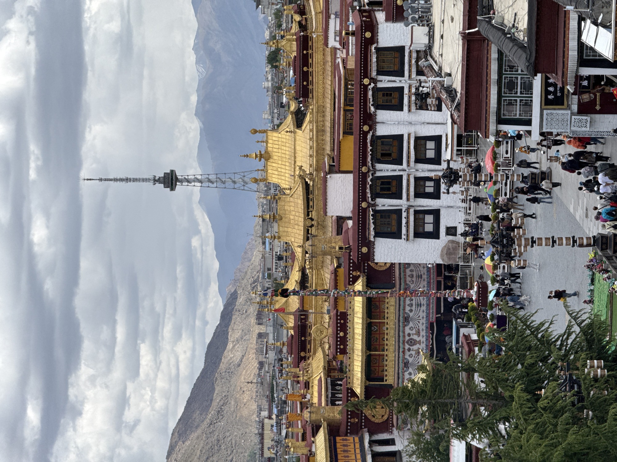 This eye-level, outdoor shot depicts the Potala Palace in Chengguan District, Lhasa, Tibet Autonomous Region, China, set against a backdrop of mountains and a cloudy sky. The palace's architecture features golden roofs and white-walled buildings with dark-framed windows. Prayer flags are strung across the area.
A crowd of people is seen walking and gathering in the courtyard. Trees are located in the lower right of the image. A radio tower is visible to the left of the Palace.