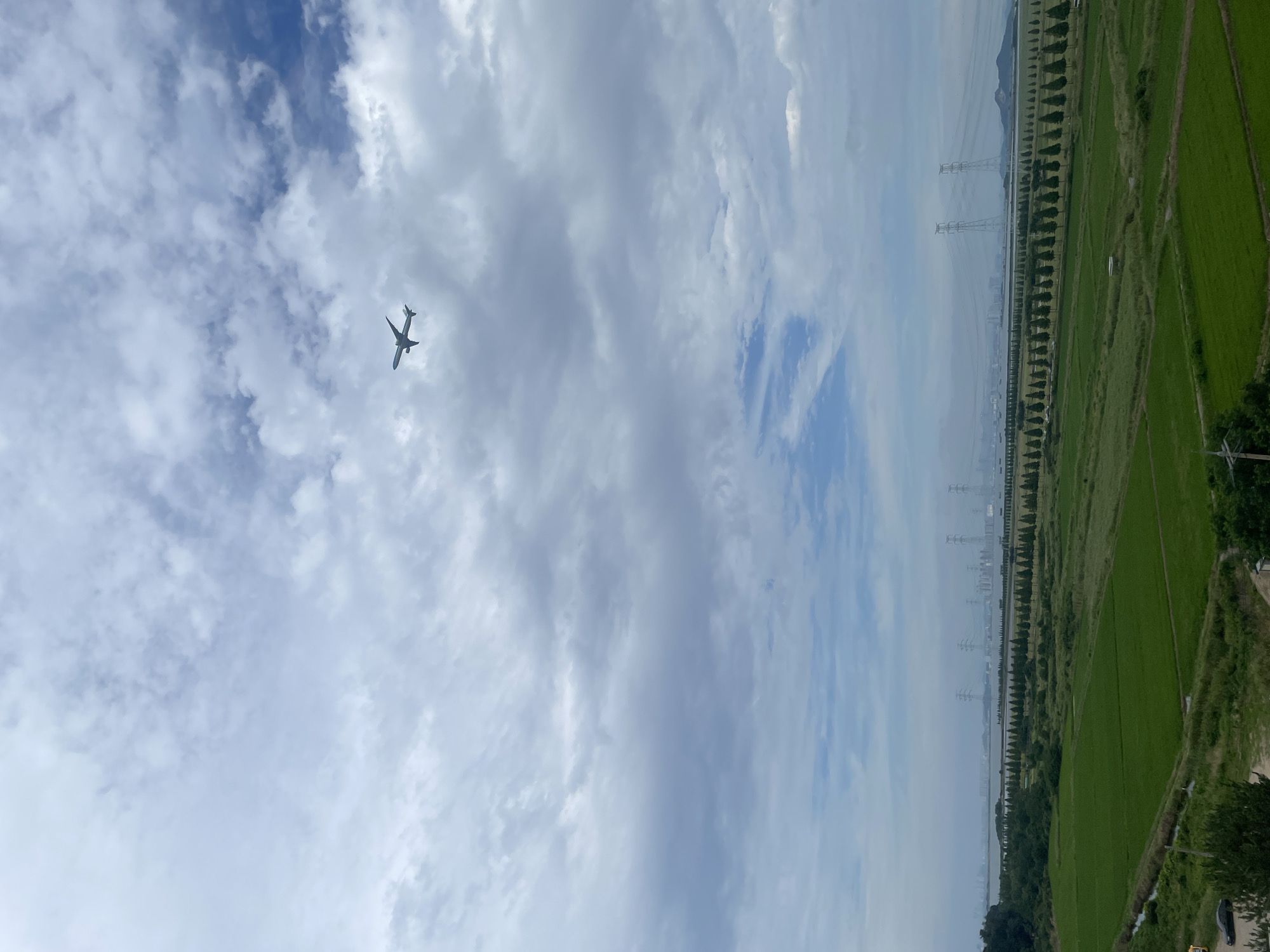 An airplane is flying in a cloudy sky above a landscape of green fields and a body of water. In the distance, there are power lines and buildings, suggesting an urban or industrial area. The scene appears to be in the city of Ansan, South Korea, likely near the coast or a large river, given the presence of the water and the flat terrain. No people are visible in the image. The overall impression is one of a vast, open sky over a developed area.