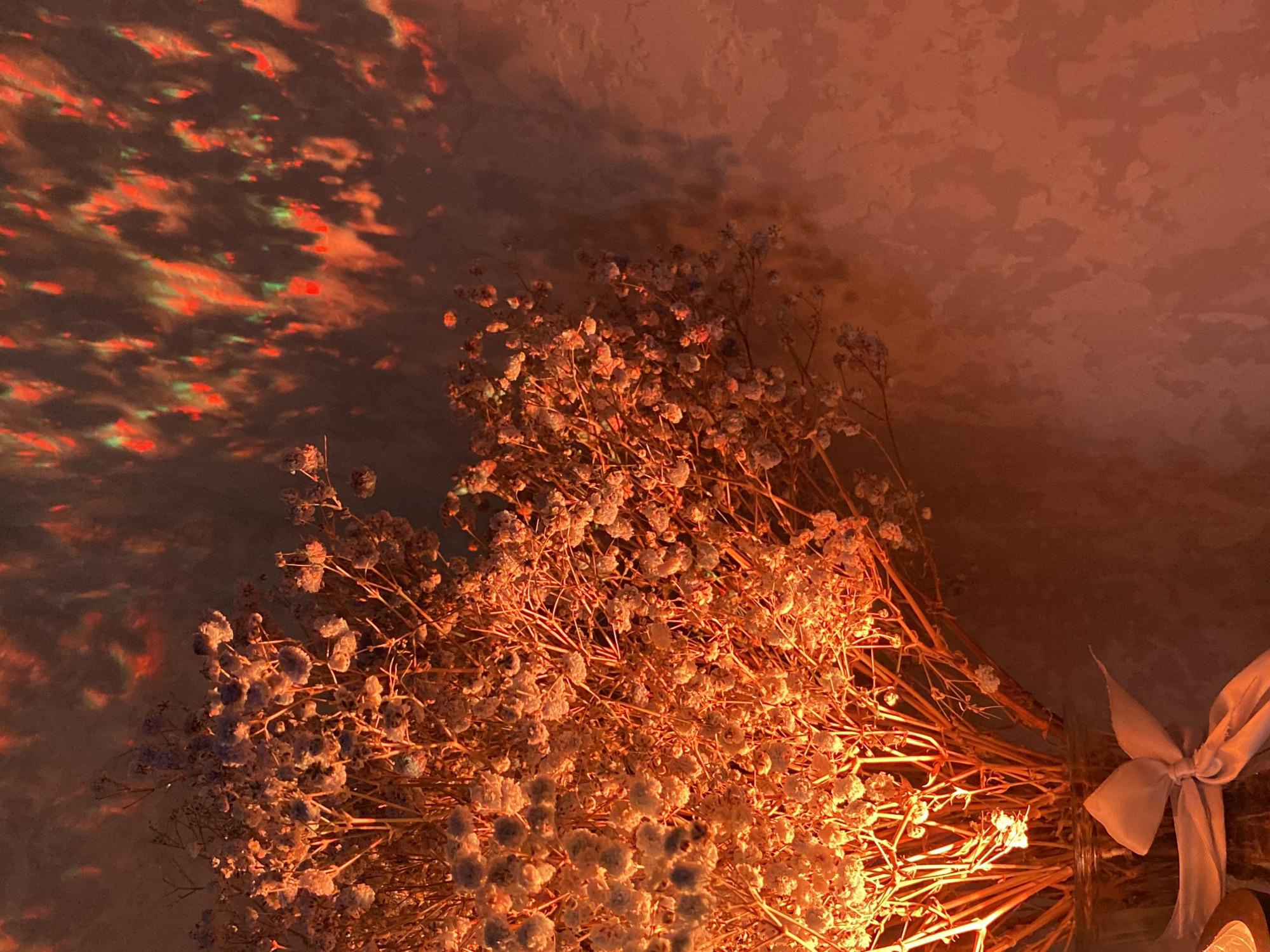 A warm-toned, atmospheric indoor photograph features a bouquet of dried baby's breath (Gypsophila) flowers under artificial lighting. The central subject is a voluminous bunch of delicate, small white or pale blue-purple blossoms with slender, dried brown stems. A light-colored, likely off-white or beige, ribbon is tied in a bow around the base of the bouquet, indicating it is likely placed in a vase or container to the lower right, partially out of frame.
Intense, warm orange and red light bathes the flowers, strongly illuminating their intricate structure and casting a bright glow, suggesting a primary light source from the upper left. The background consists of a textured wall or ceiling, also illuminated with this warm light. On the upper left side of the background, dynamic, abstract patterns of red and green light are projected, creating an iridescent, cloud-like effect. These projected patterns offer a lively contrast to the still floral arrangement.
The scene evokes a cozy and intimate ambiance, suggesting an evening or nighttime setting within a room in Izhevsk, Russia. No visible text is present in the image.