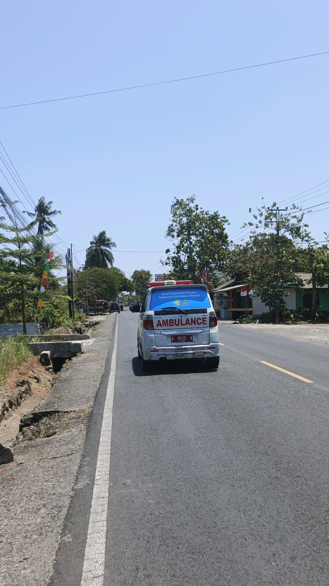 An ambulance is driving down a paved road in a rural area, likely in Desa Wonoharjo, Indonesia. The sky is clear and blue, with some overhead power lines and trees visible. The ambulance is white with a red light bar on top and "AMBULANCE" written on the back. It has a license plate that reads "Z 9975 W". There are buildings and vegetation lining the sides of the road. The road has a yellow line down the center and a white line on the shoulder. The overall scene appears to be a bright, sunny day.