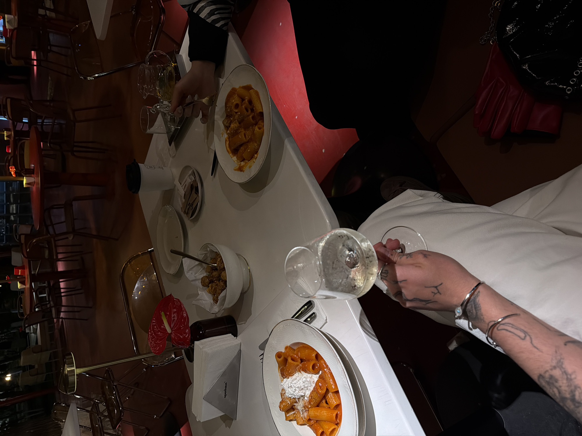 Two individuals are seated at a white table in what appears to be a dimly lit restaurant or cafe, likely in the evening. The main activity is dining and socializing, with various dishes and drinks on the table.
On the table, there are two plates of pasta, appearing to be rigatoni or paccheri, served with a rich red sauce and topped with a dollop of white cheese, possibly ricotta, and grated cheese. One person, positioned on the left side of the frame, is seen holding a fork near their pasta plate. Another person, on the right side, holds a glass of white wine. This individual's arm is visible, featuring several tattoos on the hand and forearm, and a silver bracelet with a light blue bead.
Other items on the table include a white bowl filled with what looks like small fried snacks or croquettes, a white disposable coffee cup with a black lid, and several empty glasses. A red anthurium flower adds a decorative touch, alongside a brown bottle (possibly wine or olive oil). Napkins are neatly presented in a metallic holder that reads "openhaus".
In the background, more tables and dark brown chairs are visible, suggesting an indoor dining establishment. The lighting is low, creating an intimate atmosphere, with some red accents from the background furniture. A black bag with metallic chain details and red gloves are placed on a chair or bench beside one of the diners. The scene captures a moment during a meal at a contemporary eatery in Yerevan, Armenia.