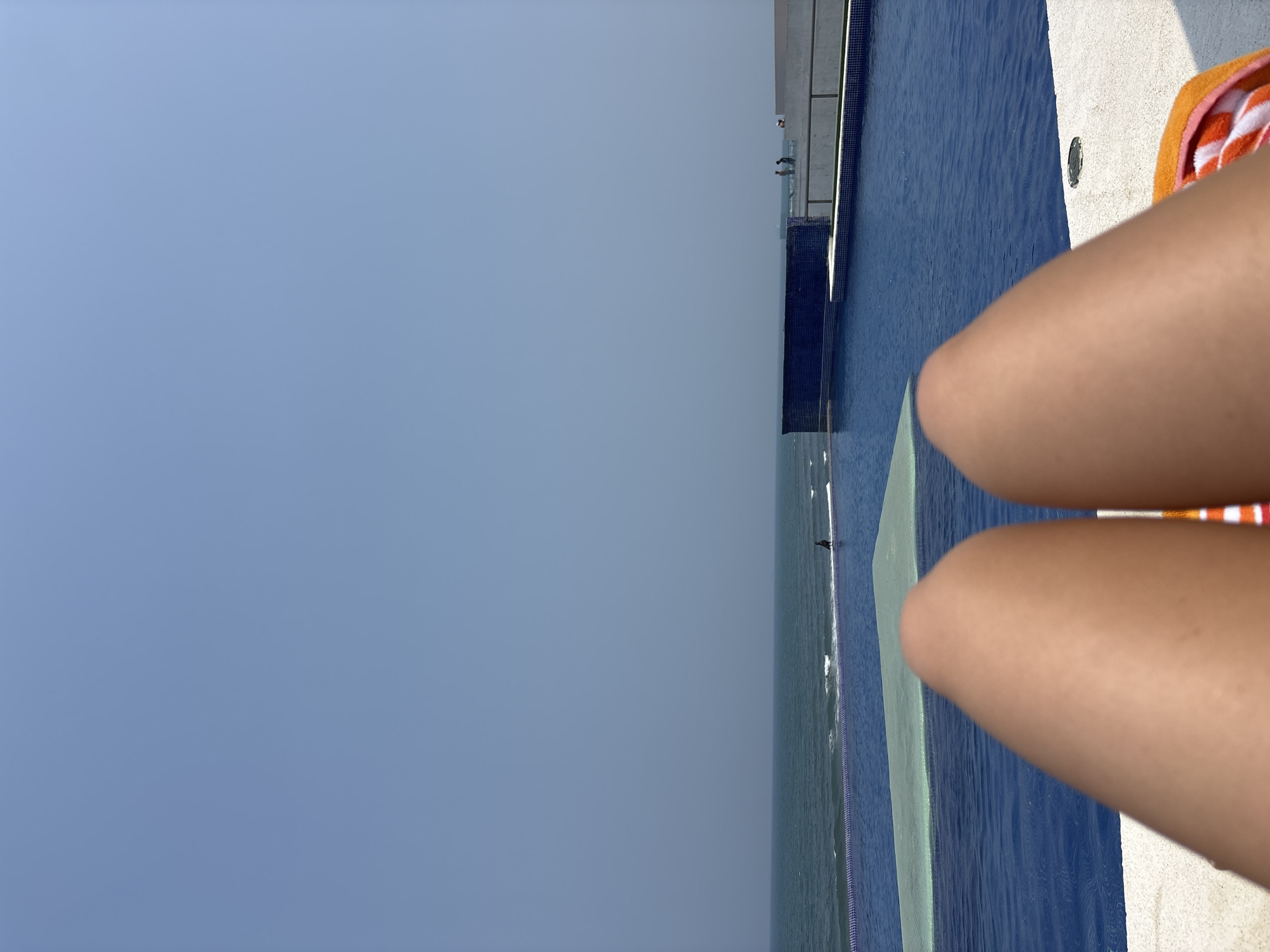 The image, taken in Boca del Río, Mexico, shows a view of an infinity pool merging with the ocean under a clear, light blue sky. In the foreground, a person's legs are visible, with an orange and white striped towel nearby. The pool's edge is tiled in dark blue. The pool appears to blend seamlessly with the sea in the background.