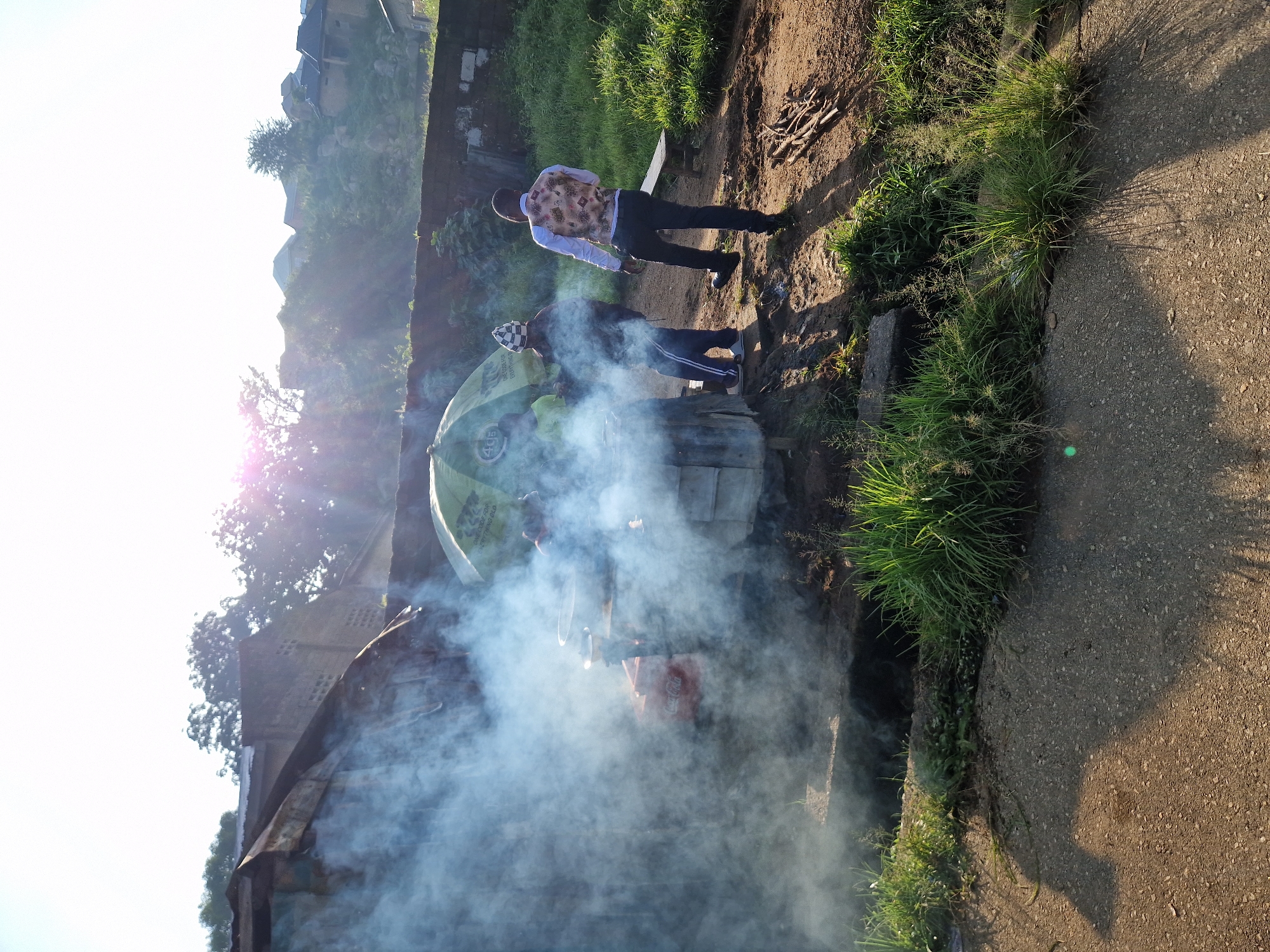 The image depicts an outdoor scene in Kabong, Nigeria, characterized by a bright, hazy sky and abundant sunlight. In the foreground, a paved surface with a rough texture is visible, casting long shadows. To the left, a substantial amount of white smoke billows from a makeshift cooking area, partially obscuring a red container with "Coca-Cola" branding and what appear to be cooking pots.
A large, green umbrella, possibly advertising "4G" and bearing the text "ONE CODE FOR EVERYTHING BILD," is positioned over the cooking area, providing some shade. Three individuals are present in the scene. One, wearing a checkered hat and dark clothing, appears to be engaged with the cooking setup. Another, standing to the right and facing away from the camera, is dressed in a white shirt and a patterned vest, with dark trousers. A third individual, also facing away from the camera, is partially visible behind the umbrella.
The environment surrounding the cooking area is unpaved, with patches of grass and dirt. A low brick wall runs along the background, behind which are buildings and trees. The overall impression is of a busy, possibly commercial, outdoor cooking operation in a semi-urban setting.