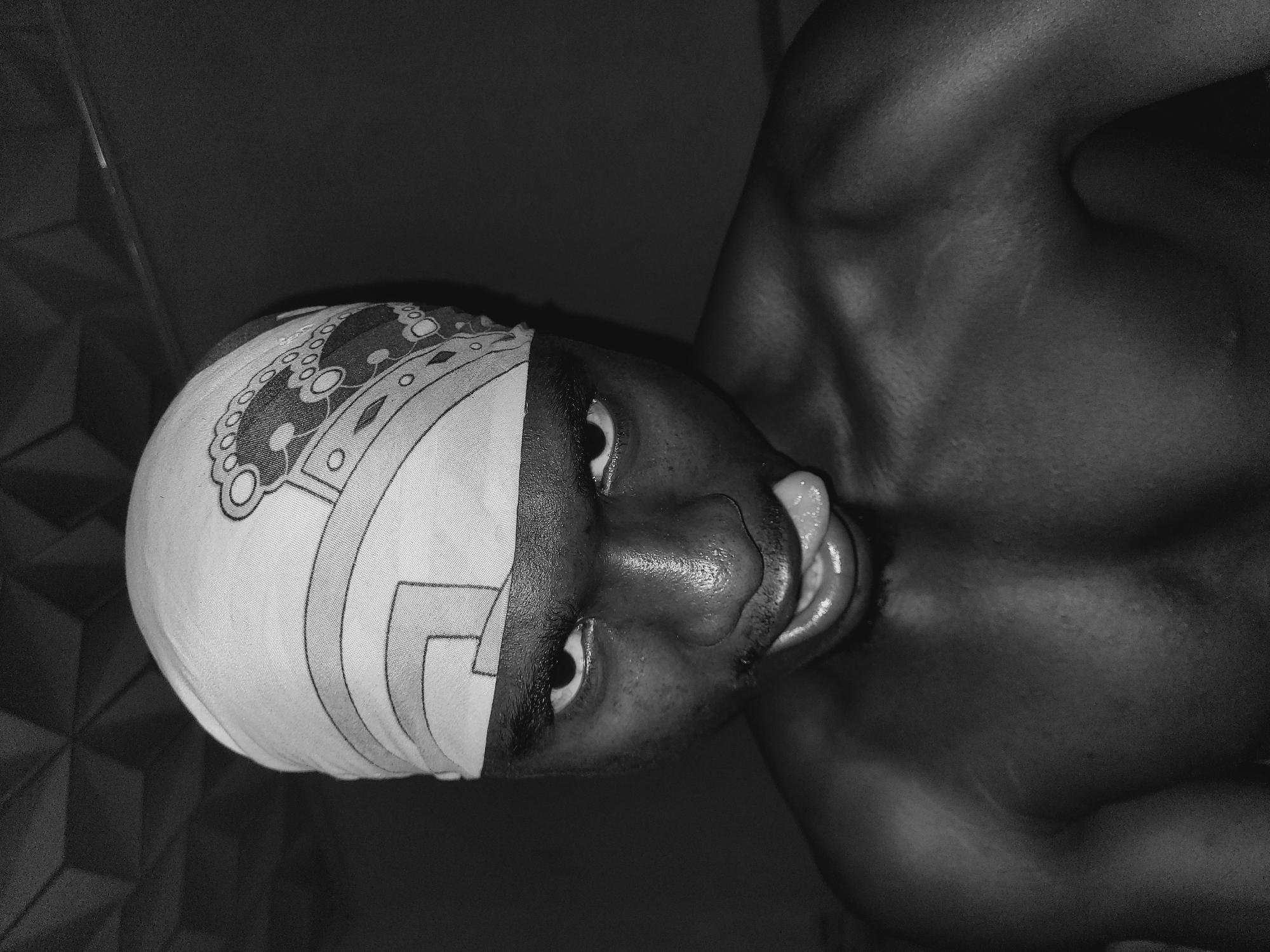 The black and white, close-up, waist-up shot shows a Black individual wearing a bandana with a patterned design, looking directly at the camera with their tongue sticking out. The individual's shoulders and upper chest are visible, and their skin appears to have some texture and minor blemishes. The background is dark and out of focus, with a patterned texture visible at the top of the frame, possibly a ceiling. The lighting highlights the individual's face and shoulders, creating a dramatic effect. There is no indication of the location in Bukuru, Nigeria.