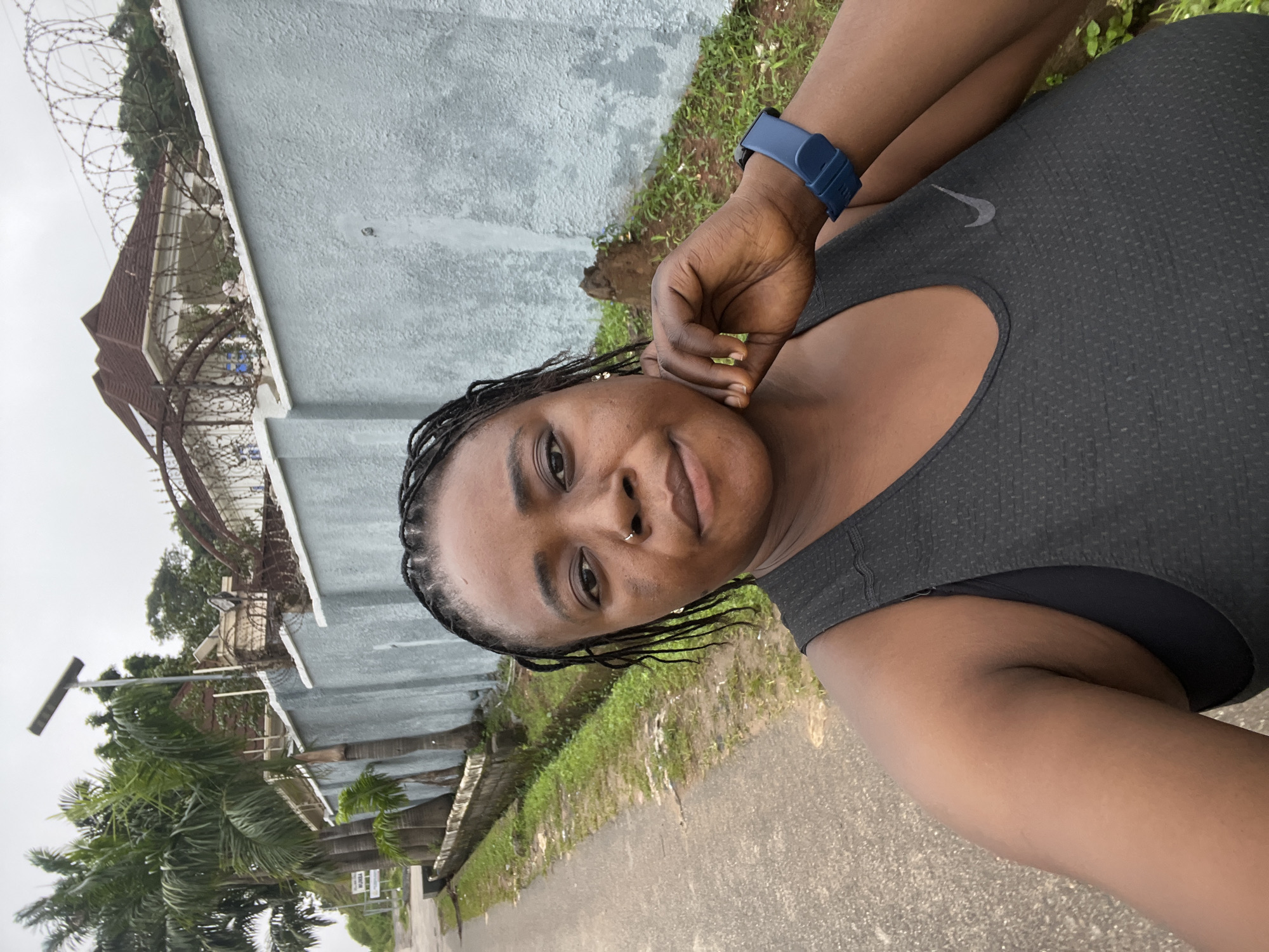 A close-up, selfie-style shot captures a Black woman in a dark grey Nike tank top, standing on a paved road with a grassy verge and a light blue wall in the background. The woman has dark braided hair, a nose ring, and is resting her chin on her right hand. She is wearing a blue wristband on her left wrist. Behind her, the wall extends to the right, topped with barbed wire. Beyond the wall, a multi-story house with a brown roof is visible, along with palm trees to the left. A solar-powered street lamp stands near the palm trees. The scene appears to be in an urban or suburban residential area, possibly in Abuja, Nigeria, given the architecture and vegetation. The lighting suggests an overcast day.