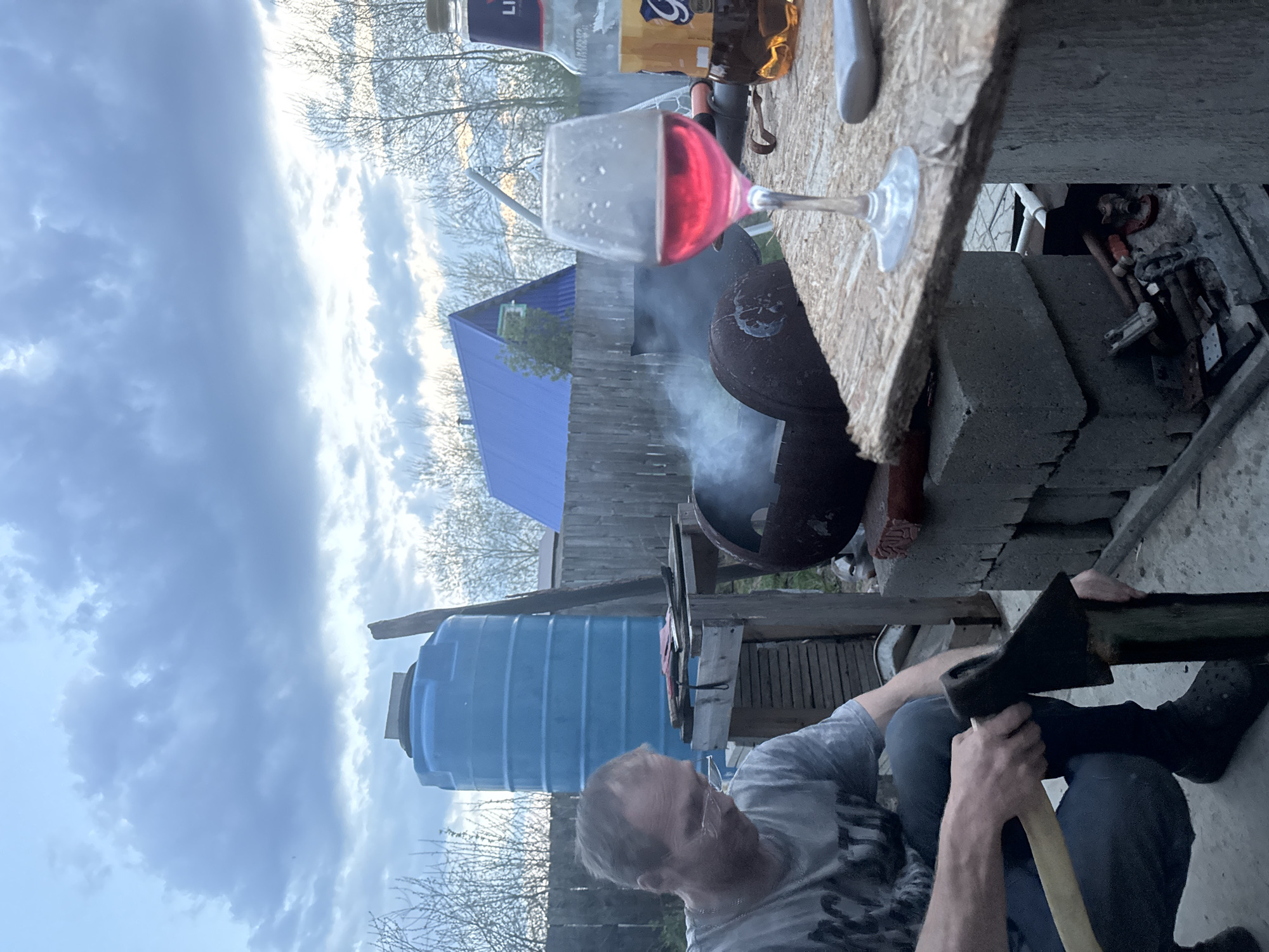 The image depicts an outdoor scene in what appears to be a backyard setting in Chaikovsky, Russia. A man is seated, holding an axe. Behind him, there is a blue water tank and a partially obscured blue-roofed structure. Smoke rises from a grill or outdoor cooking device. On a table near the grill, there is a glass of red liquid, possibly wine, and bottles of alcoholic beverages. The environment suggests a rural or semi-rural area with fences and trees visible in the background. The sky is cloudy.