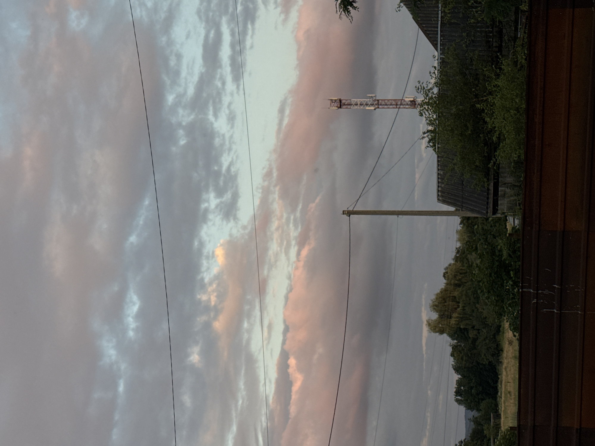 In the city of Zassovskaya, Russia, a late afternoon or early evening scene unfolds. The sky is filled with a dramatic display of clouds, tinged with soft pinks and grays, indicating the approaching sunset. Power lines crisscross the frame, adding a man-made element to the natural landscape.
Dominating the middle ground is a tall, red and white telecommunications tower, a modern structure piercing the sky. To the right of the tower, the corrugated roof of a building is visible, partially obscured by lush green foliage. To the left of the tower, a wooden utility pole stands, further emphasizing the presence of infrastructure.
In the background, a dense line of trees forms a natural border, with a field of dry grass stretching out before them. The foreground is marked by a rusty, weathered metal barrier, perhaps a fence or industrial debris, with white splatters, possibly paint or bird droppings, adorning its surface. The overall impression is one of rural quietude, with a blend of natural and man-made elements under a brooding sky.